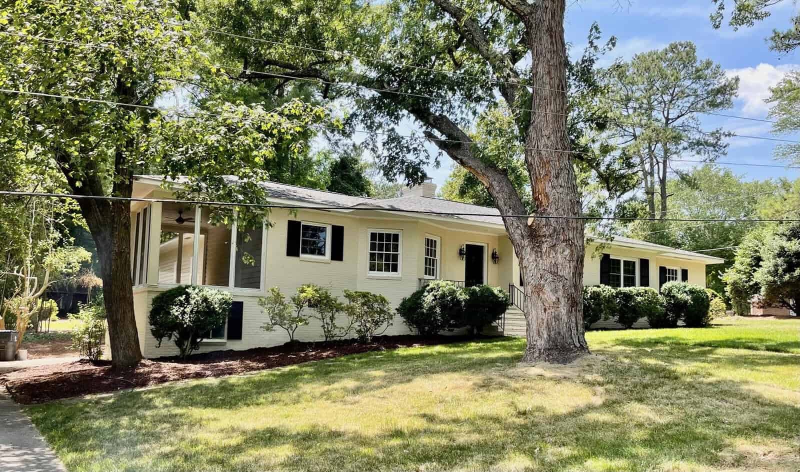 A single-story beige house with a spacious front yard featuring a large tree and well-maintained shrubs. The house has multiple windows, a small front porch with a railing, and a walkway leading to the entrance. There are tall trees and a blue sky in the background.