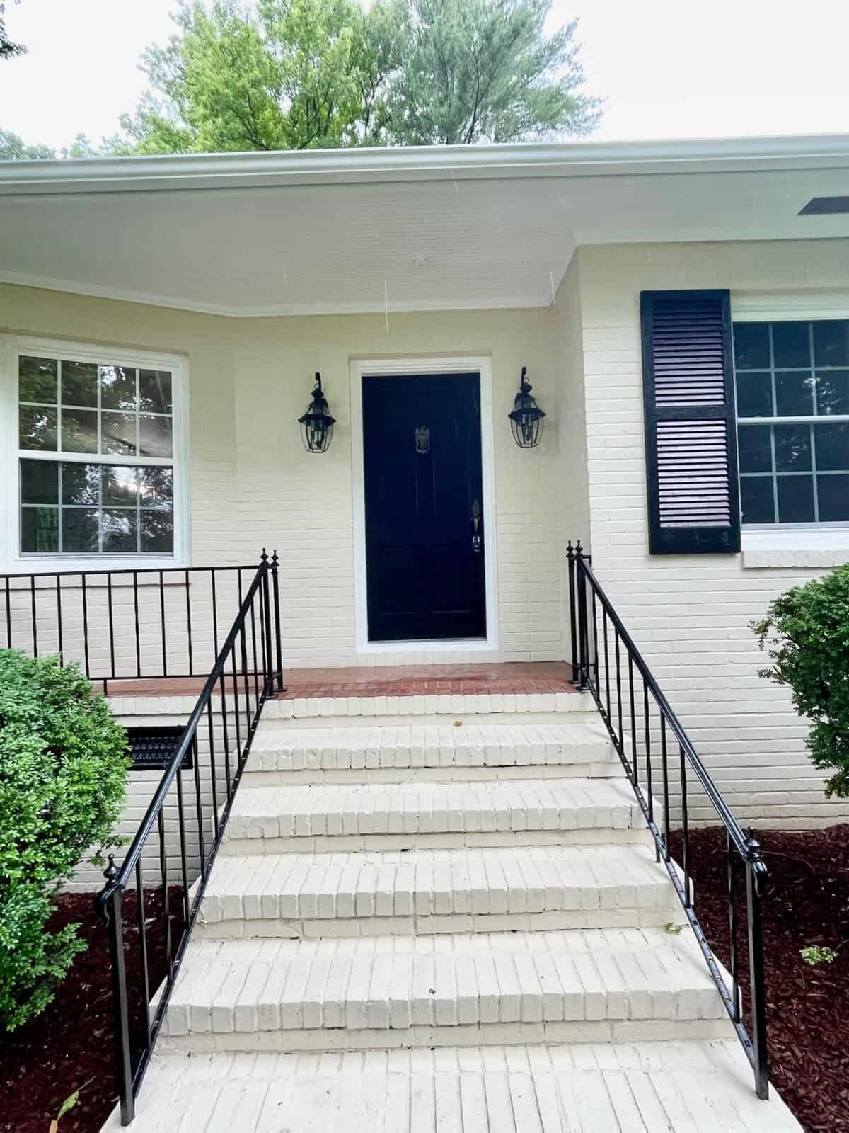 A front entrance of a house featuring a dark blue door, flanked by two windows with black shutters. Six white brick steps with black iron railings lead up to the door. Two black lantern-style lights are mounted on either side of the entrance. Shrubs line both sides of the steps.