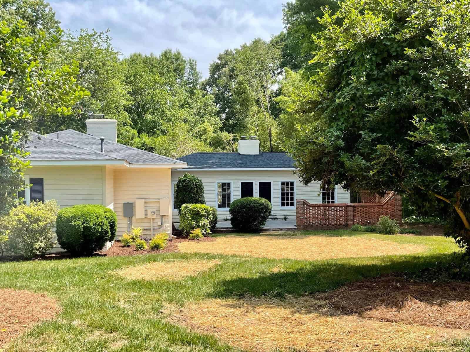 A charming single-story house with light-colored siding and a dark gray roof, surrounded by lush green trees and bushes. There's a wooden patio area with a lattice fence on the right side, and a neatly manicured lawn in the foreground. The sky is partly cloudy.