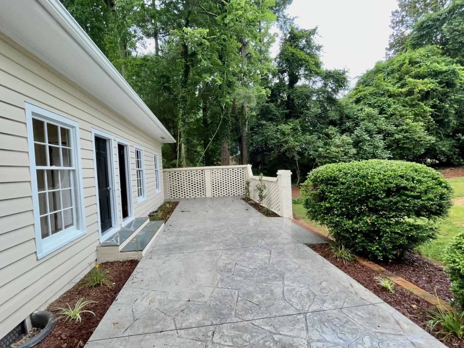 A stone walkway leads to a beige house with white trim and several windows. A white lattice fence borders the walkway, and there is greenery, including shrubs and tall trees, surrounding the area. The setting is outdoors, in a serene and lush environment.