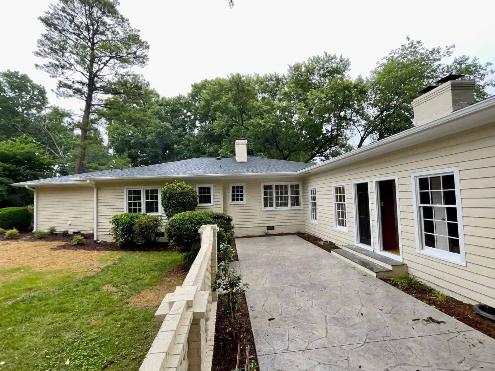 A single-story beige house with a large stone patio, multiple white-framed windows, and a brown front door. The house is surrounded by a green lawn, shrubs, and trees. A low beige brick wall borders the patio, and a tall tree stands to the left.