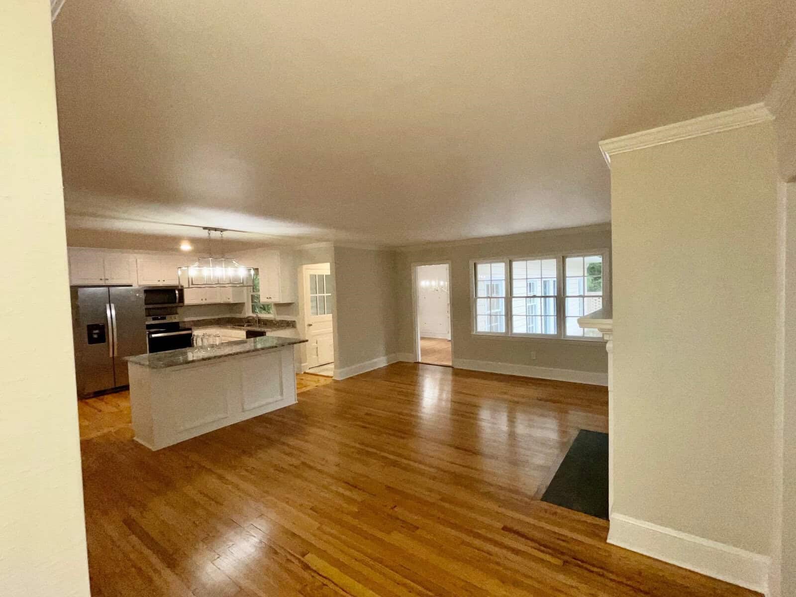 An empty, modern kitchen and living area featuring hardwood floors, white walls, and a kitchen island with a granite countertop. The kitchen includes stainless steel appliances, and large windows on the right side offer natural light to the space.