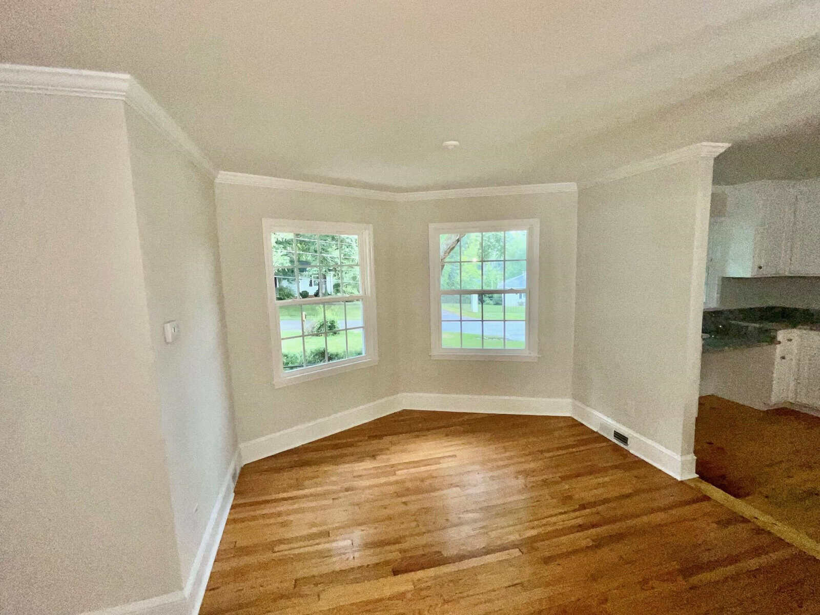 A small room with hardwood floors and light gray walls, featuring two large windows overlooking a green yard outside. The room has white ceiling trim and baseboards. The adjacent area with white cabinets and a marble countertop is partially visible.