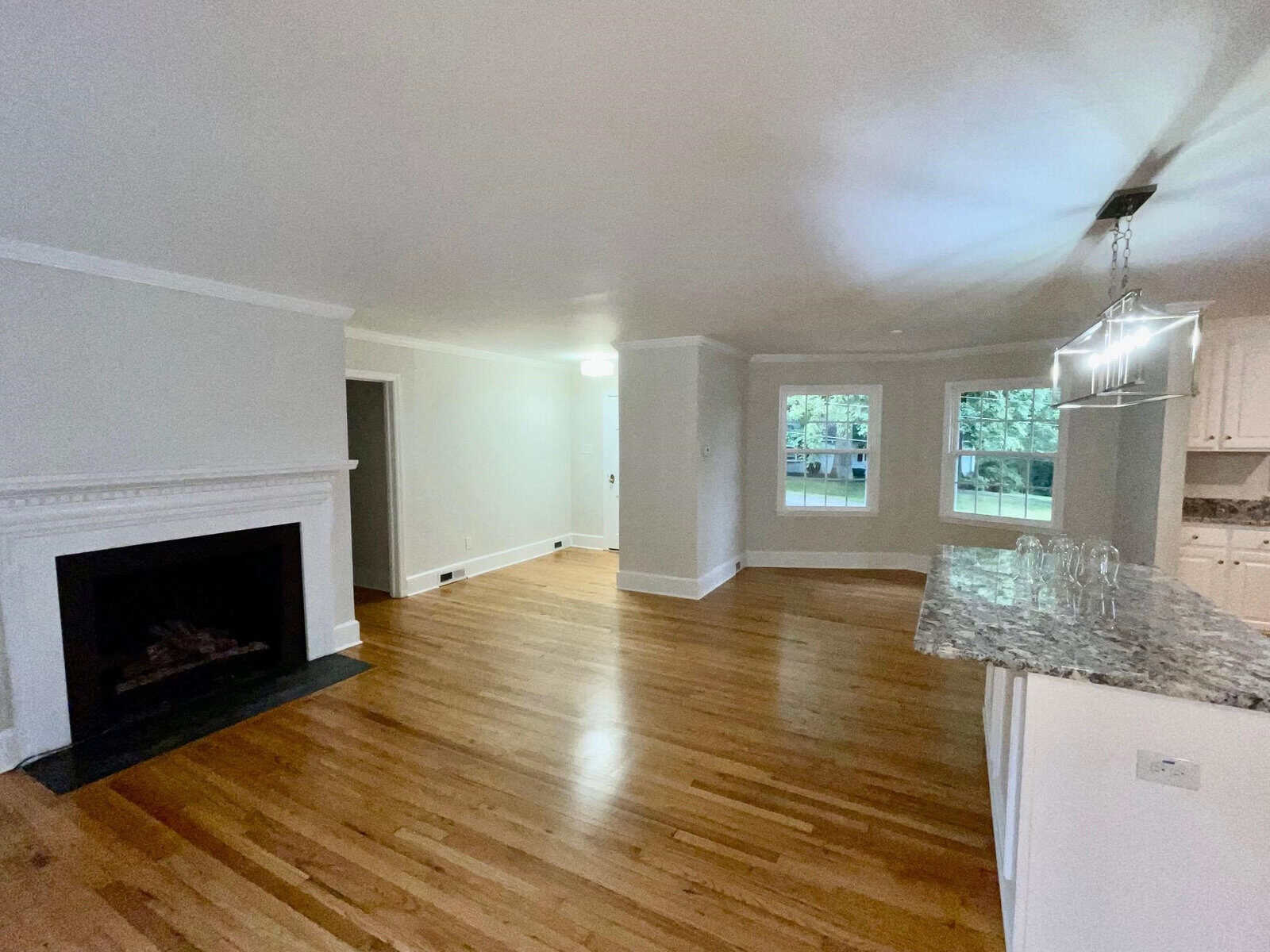 A spacious, light-filled living room with polished hardwood floors, a white fireplace on the left, and an open kitchen area with a granite countertop and hanging light fixture on the right. Large windows offer a view of the greenery outside.