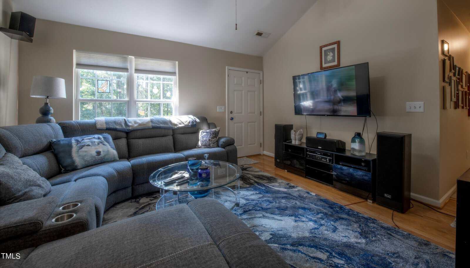 A cozy living room with a large gray sectional sofa, a glass coffee table, and a flat-screen TV mounted on the wall. Natural light streams through two windows. The room is decorated with a blue-patterned rug and a lamp in the corner.