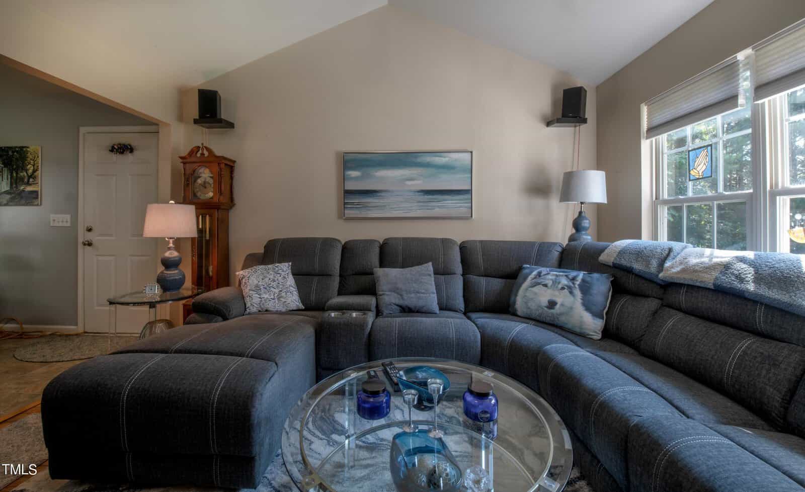 A cozy living room with a large sectional sofa, adorned with a dog-themed pillow. A round glass coffee table holds decor items, while a painting hangs on the wall above. Natural light streams through the windows, illuminating the space.