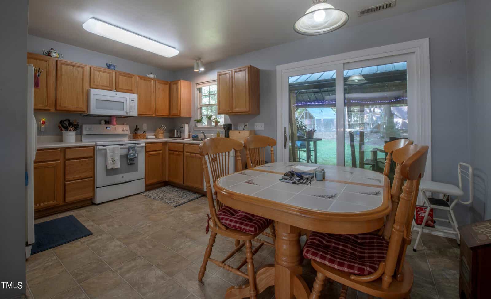 A kitchen and dining area with wooden cabinets and a tiled floor. The space features a stove, oven, and a refrigerator. A wooden dining table with four chairs is centered, and a sliding glass door opens to a patio with a view of the outdoors.