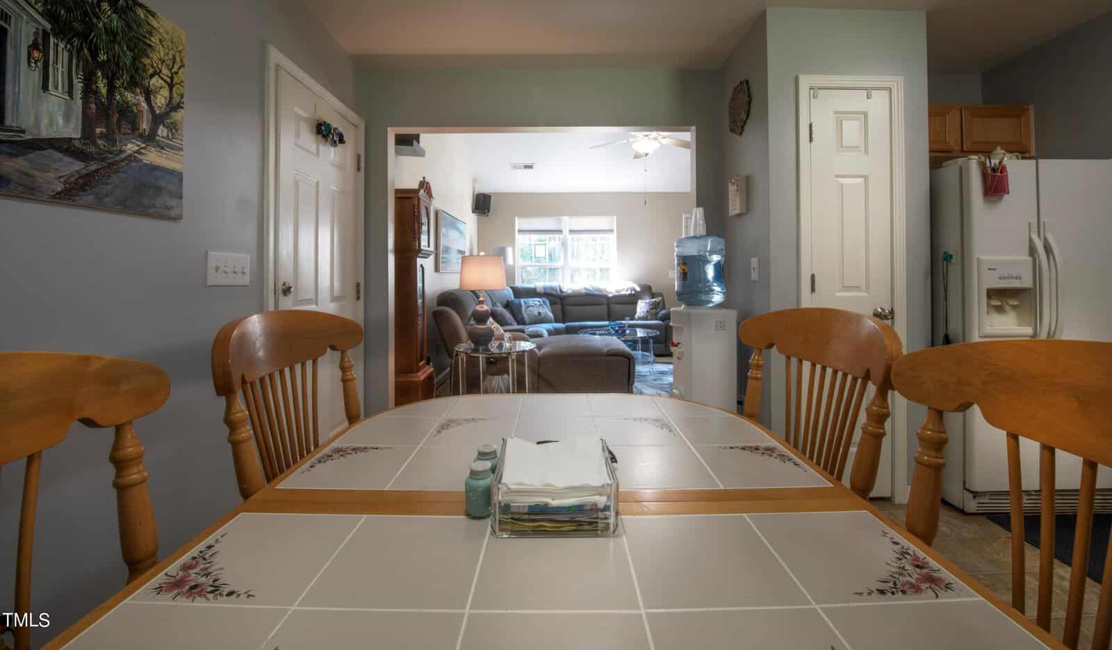 A kitchen view showing a tiled table with wooden chairs. In the background, a living room is visible with a large sofa, coffee table, ceiling fan, and windows letting in natural light. A fridge is to the right and a painting hangs on the wall to the left.