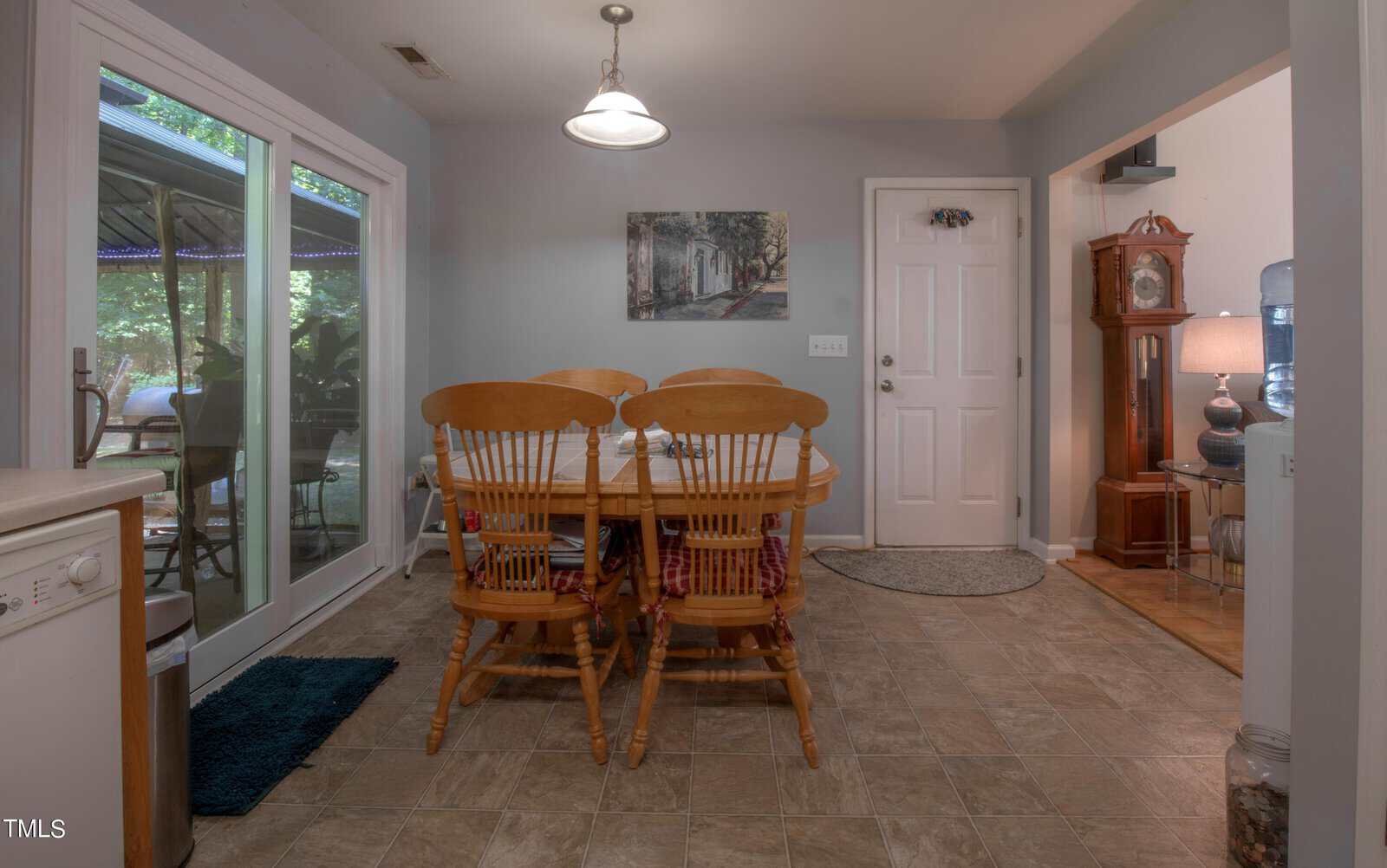A dining room features a wooden table with four chairs. There's a clock against the wall, a painting above it, and a door on the side. Large sliding glass doors open to an outdoor area, allowing natural light to fill the room.