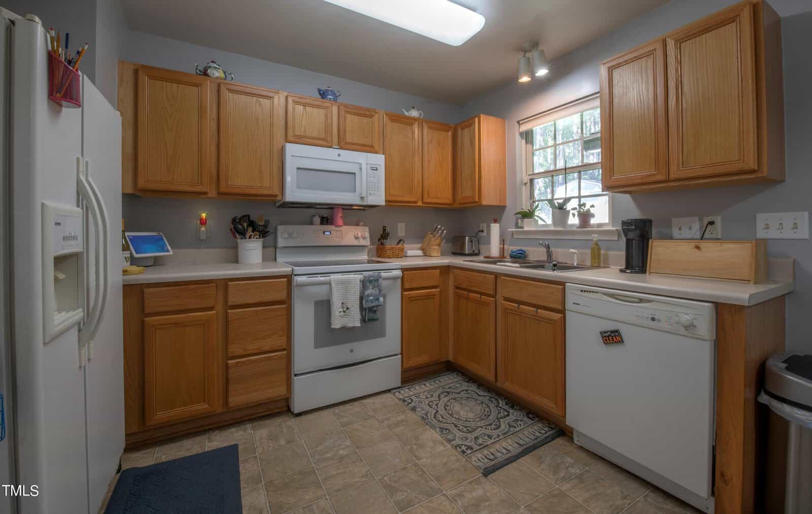 A home kitchen with wooden cabinets, white appliances, and a tile floor. There's a rug in front of the sink, and sunlight streams through a window above the counter. Various kitchen items are placed on the countertops.