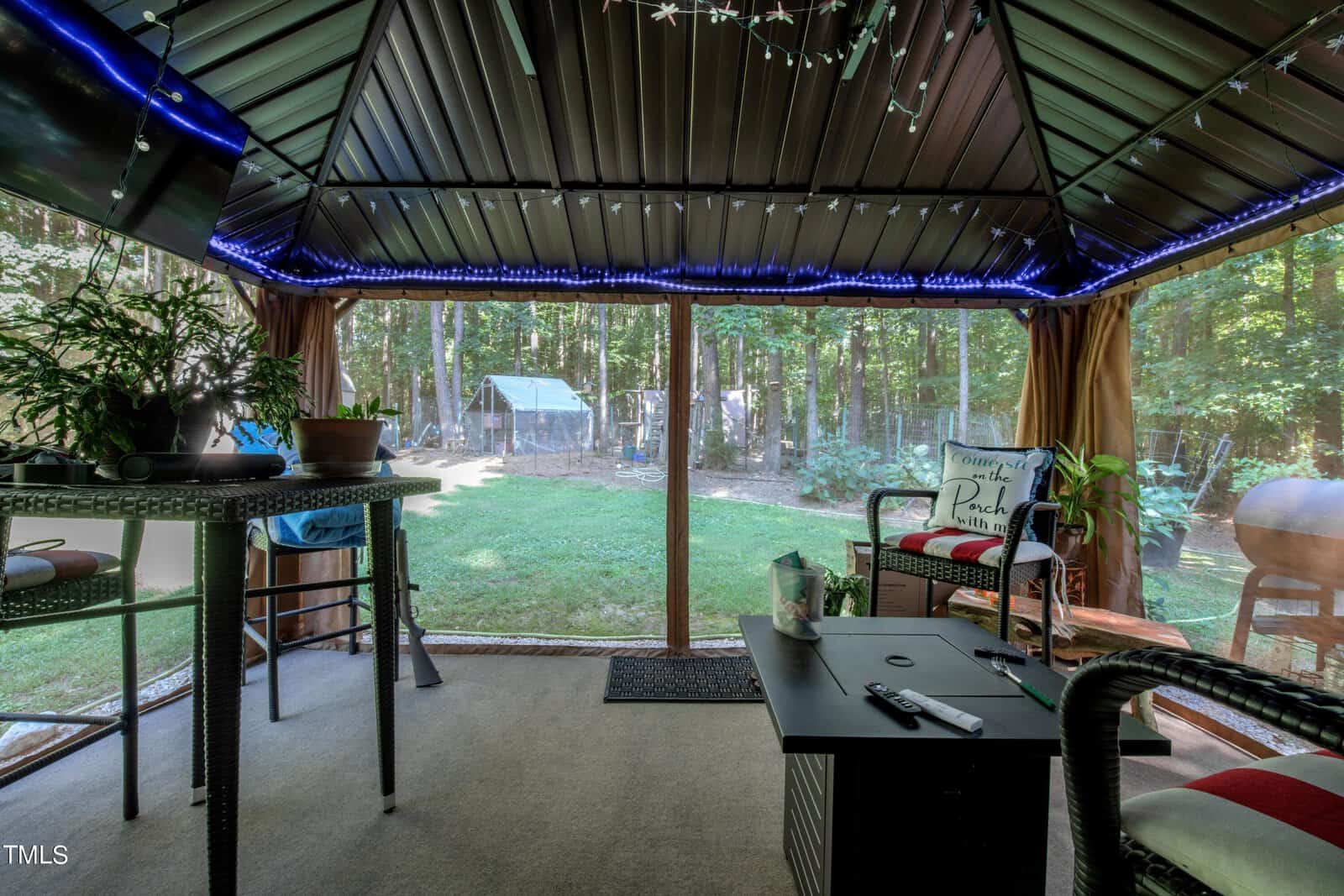 A cozy outdoor gazebo with string lights, a bar table with stools, potted plants, and a seating area with a cushion that says "Sit, Relax, Enjoy." Through the screened walls, a lush garden and a greenhouse are visible.