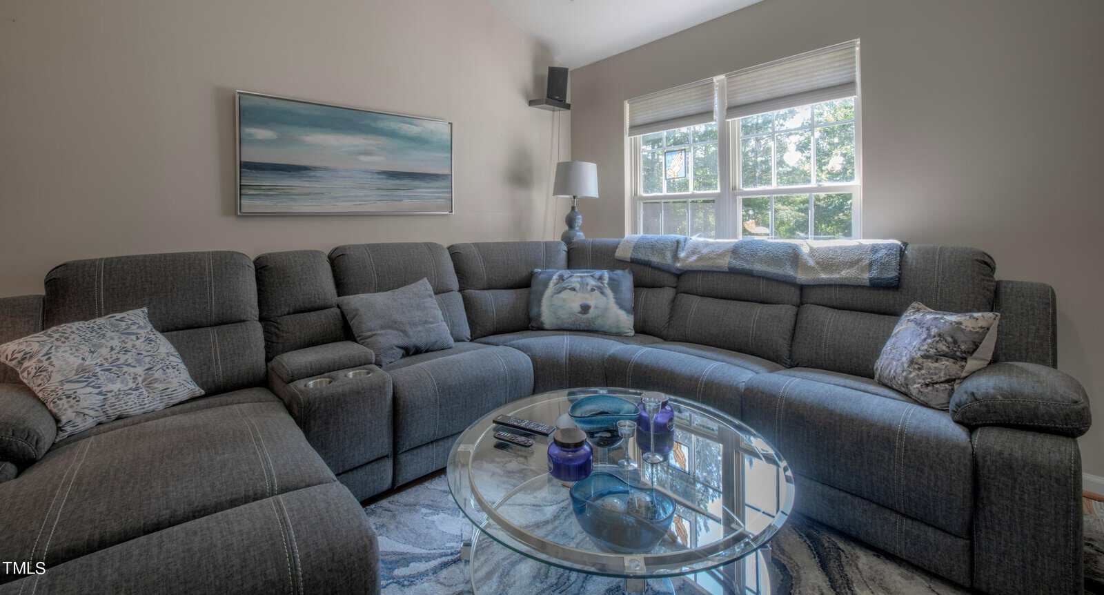 A cozy living room features a large gray sectional sofa with decorative pillows. A round glass coffee table holds various items in the center. A painting hangs on the wall, and natural light streams through the two windows, illuminating the space.
