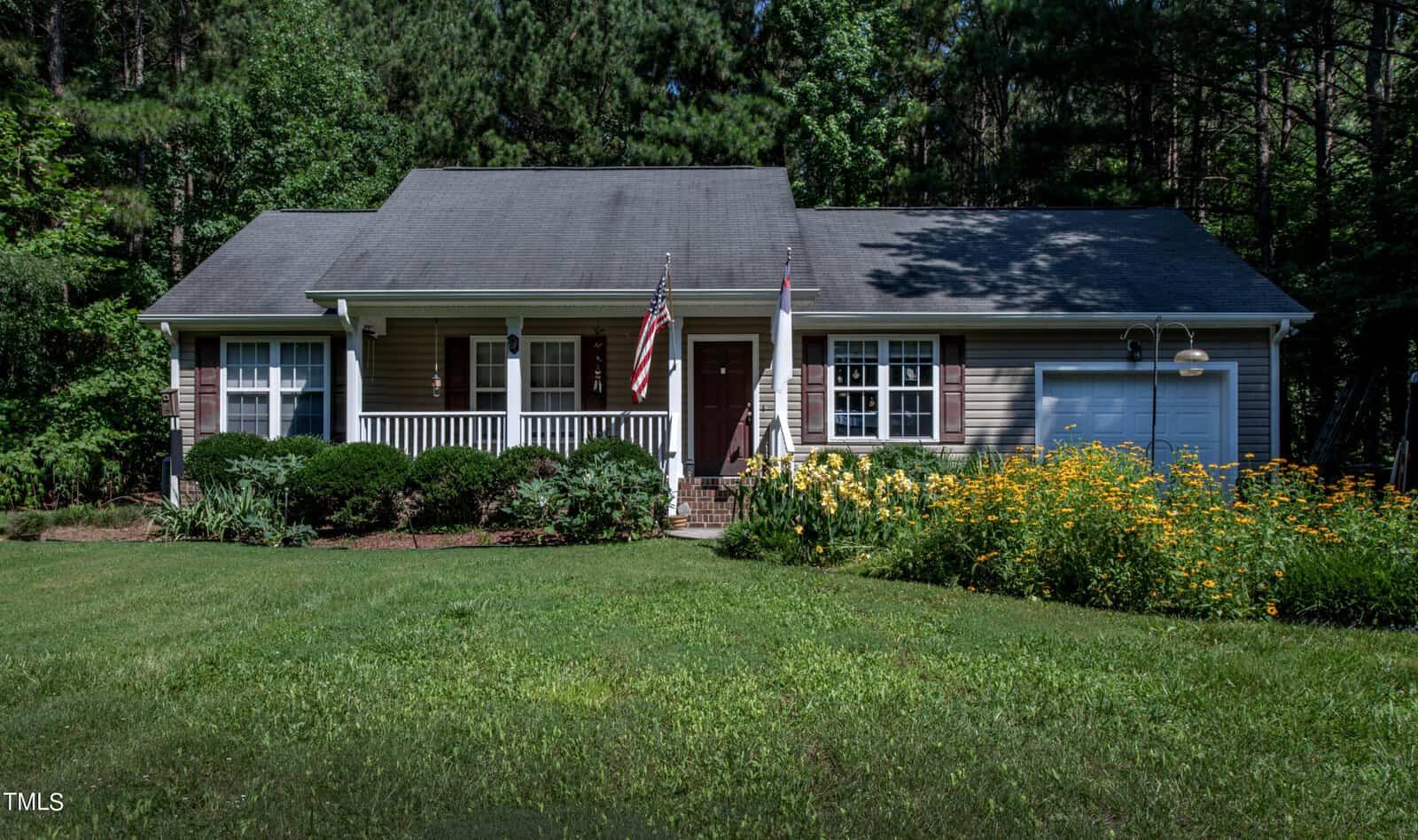 Nestled on 2125 Sterling Creek Lane in Franklinton, this single-story house boasts a porch adorned with flags and an inviting garage. Encircled by lush greenery, it features a manicured lawn, vibrant yellow flowers and shrubs. Towering trees provide a scenic backdrop, casting gentle shadows on the roof.