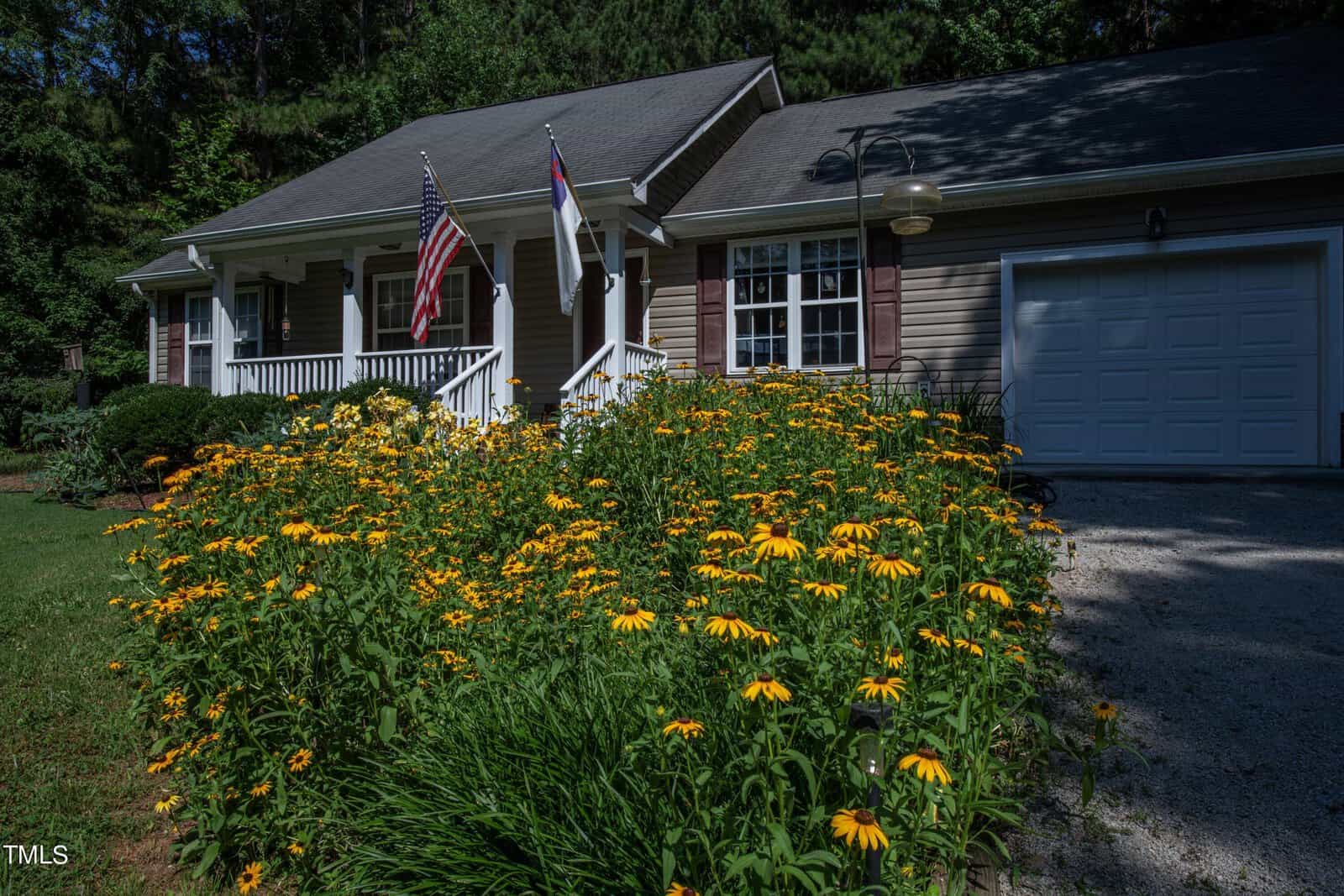 A quaint house with a front lawn filled with blooming yellow flowers. Two flags are mounted on the porch, and a white garage is on the right. The house is surrounded by lush green trees under a clear blue sky.