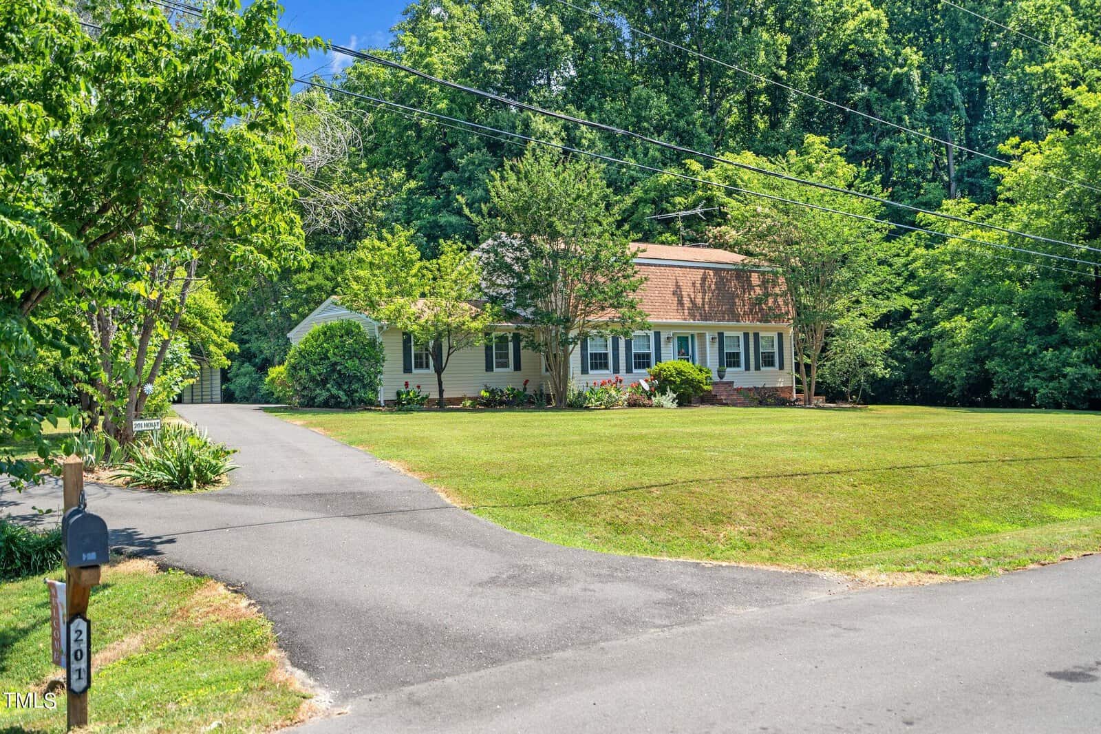 A charming suburban house with a brown roof and light-colored siding is surrounded by a well-maintained lawn and lush trees. A paved driveway leads to the house, and a mailbox labeled "20" is visible in the foreground.