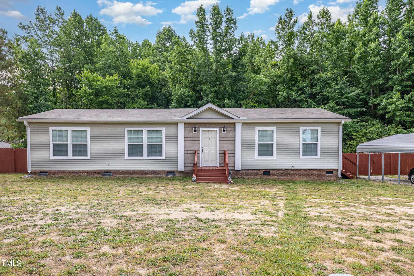 A single-story, beige manufactured home with a brick foundation and a front porch featuring brown steps. It is surrounded by a spacious yard with a backdrop of tall, leafy green trees under a partly cloudy sky.