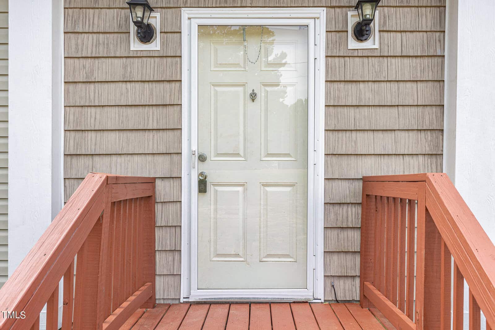 Front view of a house entrance featuring a white door with a small knocker. The door is flanked by two black lantern-style lights. A wooden ramp with red-brown railings leads up to the door. The exterior wall is clad in beige siding.
