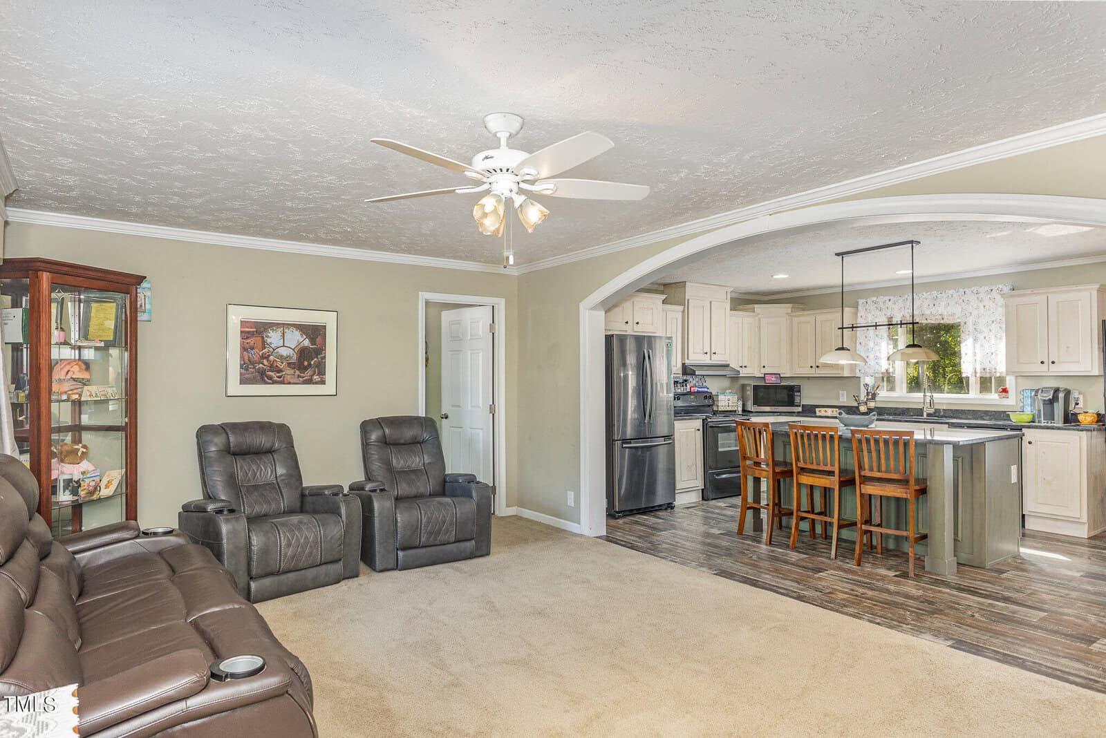 A cozy living room with two recliners facing a sofa, cream-colored walls, and a ceiling fan. The adjacent kitchen features a breakfast bar with wooden stools, stainless steel appliances, and white cabinets. A display cabinet and artwork are on the left wall.
