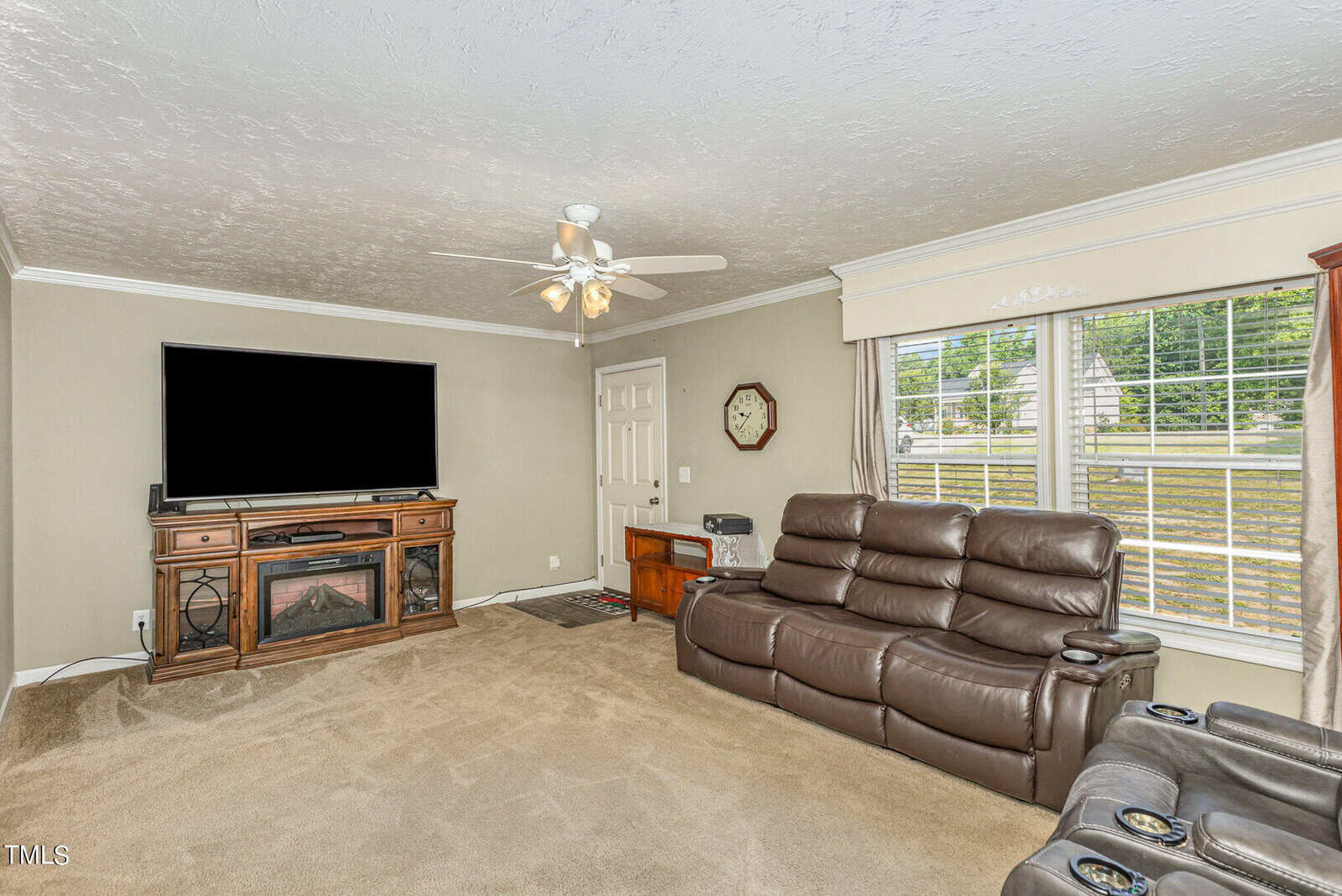 A cozy living room with a large, wall-mounted TV and a wooden console. There's a brown leather reclining sofa facing the TV. Large windows with blinds let in light, and a ceiling fan with lights is above. A door and clock are on the back wall.