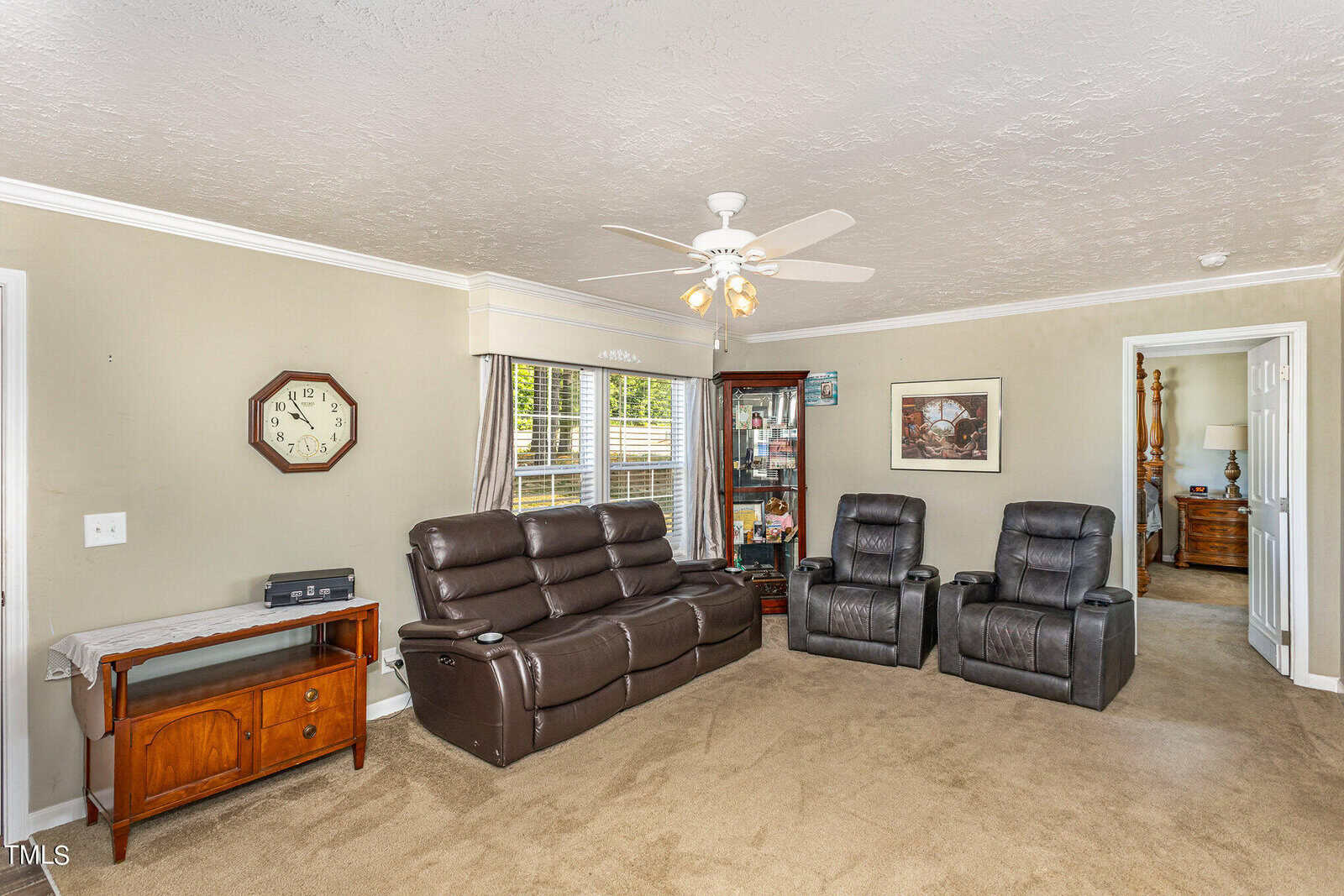 A cozy living room with beige walls and carpet features a brown leather sofa and two matching recliners. A ceiling fan hangs above, and a wooden cabinet with glass doors stands by the window. A wall clock and small wooden table complete the decor.