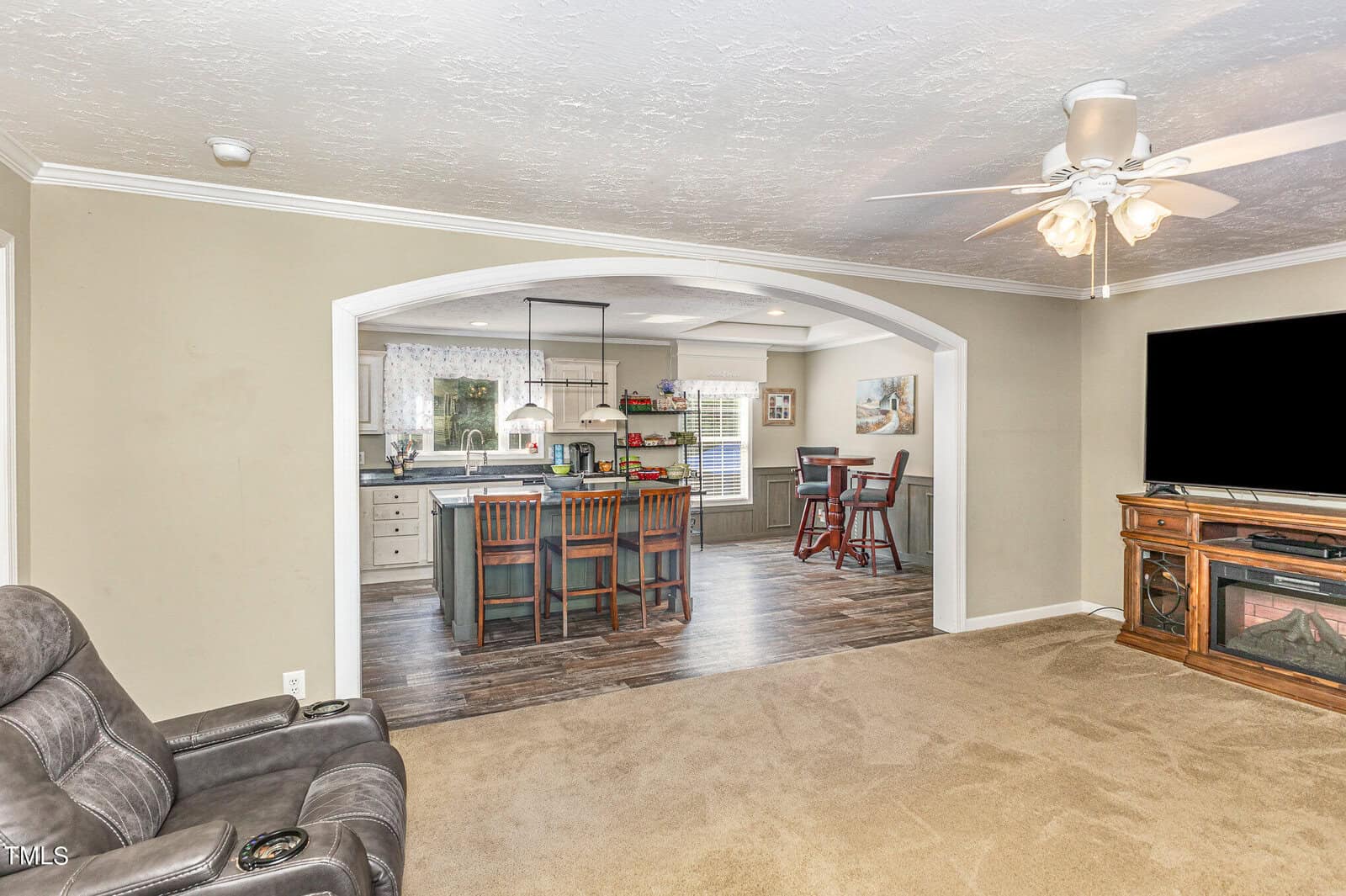 Spacious living area with a ceiling fan, armchair, and large TV. Adjacent is an open-plan kitchen featuring a kitchen island with bar stools and dining area. Wood flooring contrasts with neutral carpet and light walls. Natural light filters through blinds.