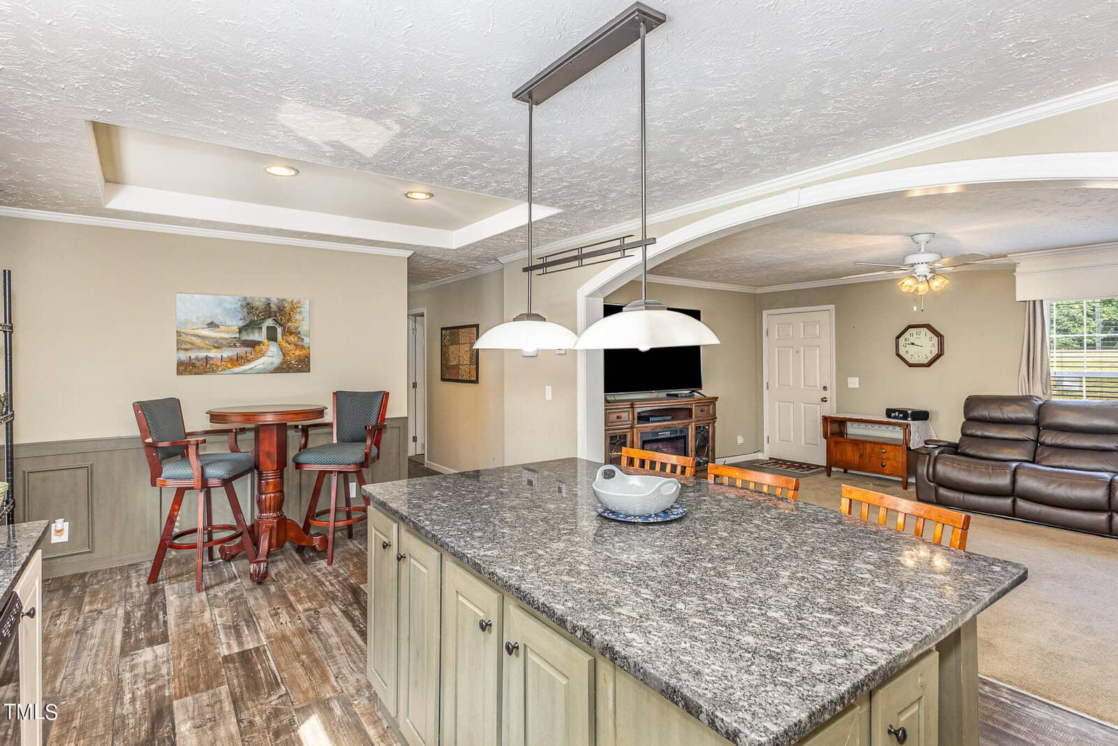 A modern kitchen with a granite island and wooden bar stools. A living area in the background features leather recliners, a TV, and a ceiling fan. Recessed lighting highlights the space, and a painting decorates the wall.