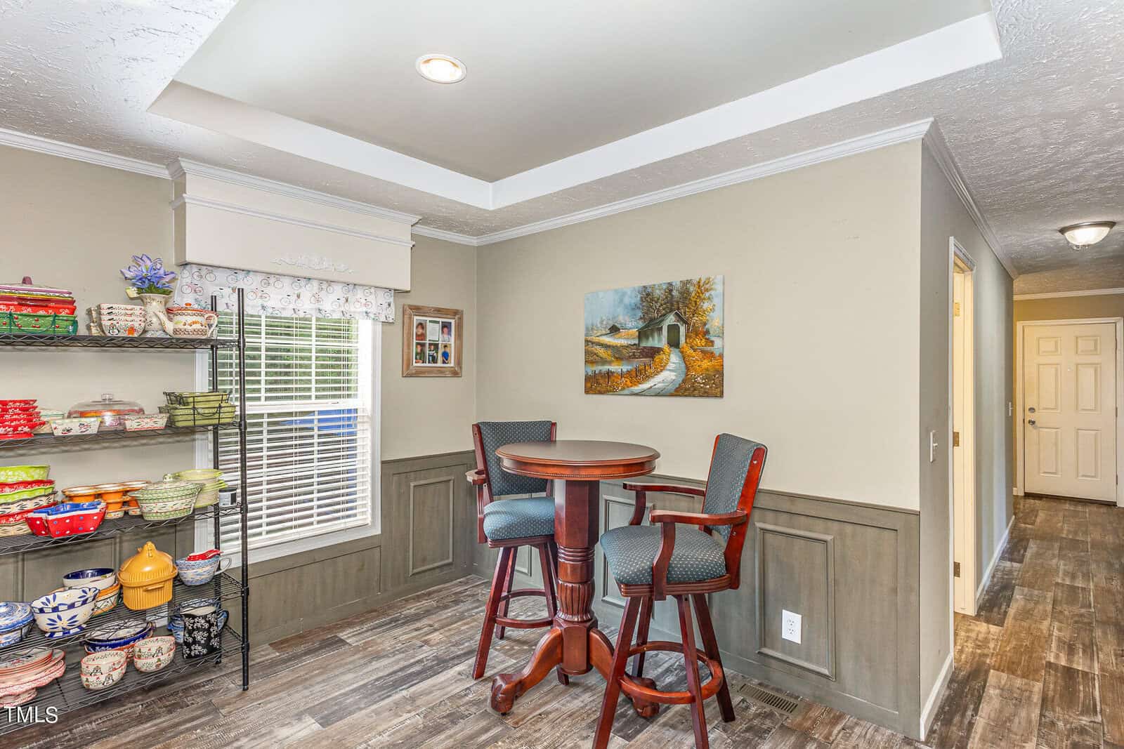 A cozy dining area with a tall round table and two chairs. A shelf filled with colorful dishes is on the left. A window with a valance lets in light, and a painting of a path through trees hangs on the opposite wall. Wood flooring adds warmth.
