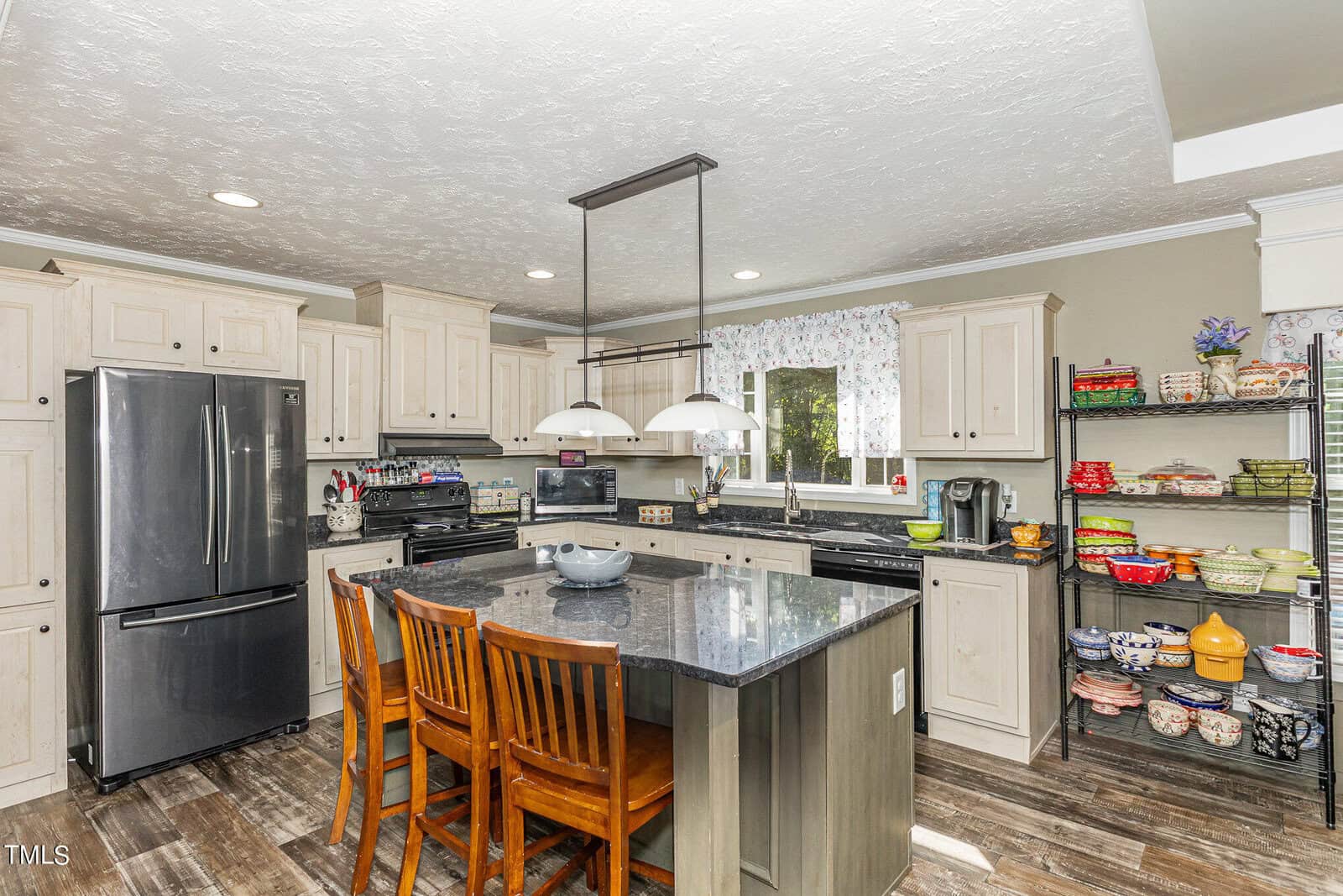 A modern kitchen with light wood cabinets and a gray island with a dark countertop. Three wooden chairs surround the island. Stainless steel appliances are present, along with open shelving filled with colorful dishes and items. A window is above the sink.