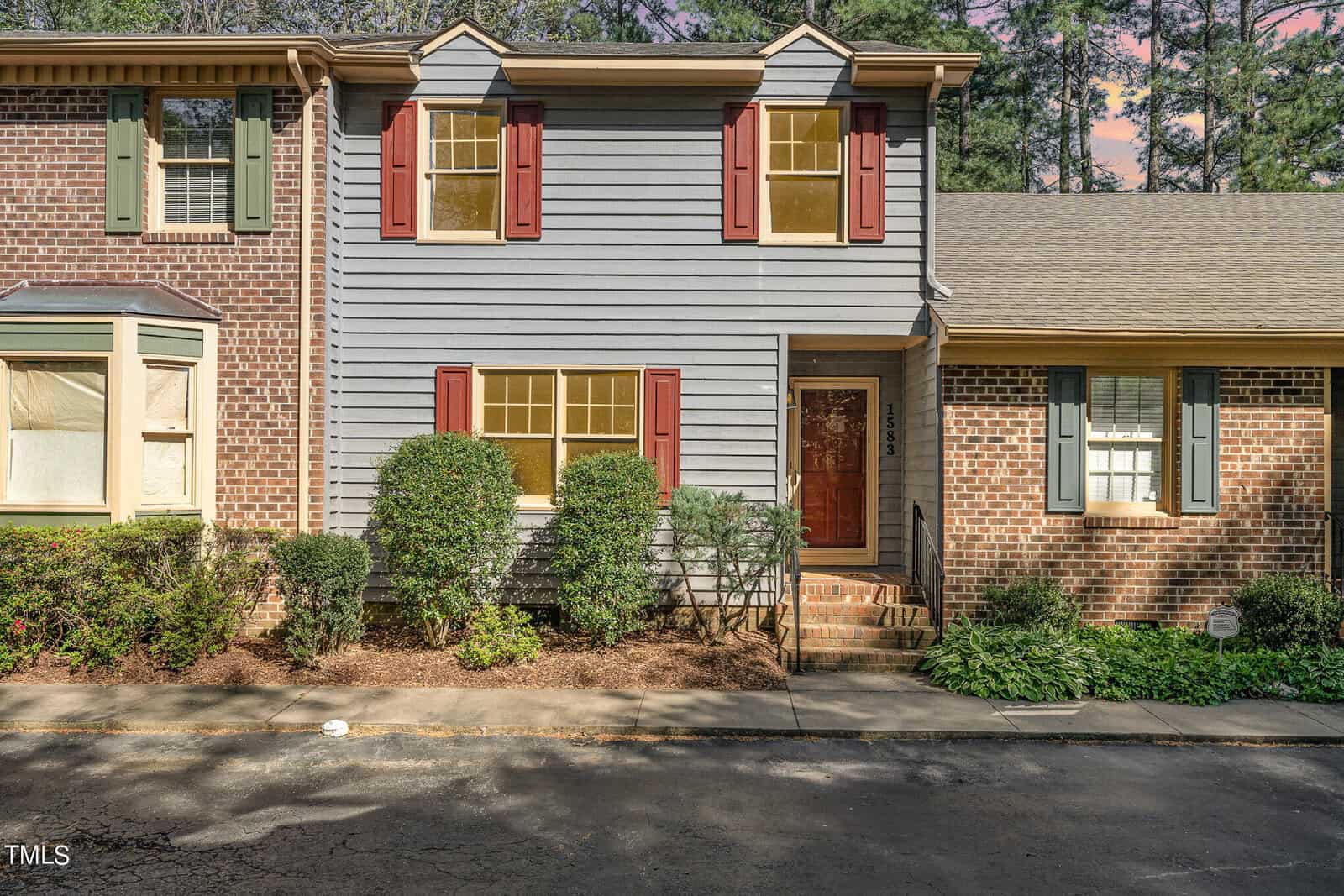 A two-story townhouse at 1583 Graham Avenue, Henderson NC, features blue-gray siding and red shutters, flanked by brick facades. The entrance is reached by a small set of steps, and the property is surrounded by bushes. Trees and a pinkish sky are visible in the background, indicating sunset.