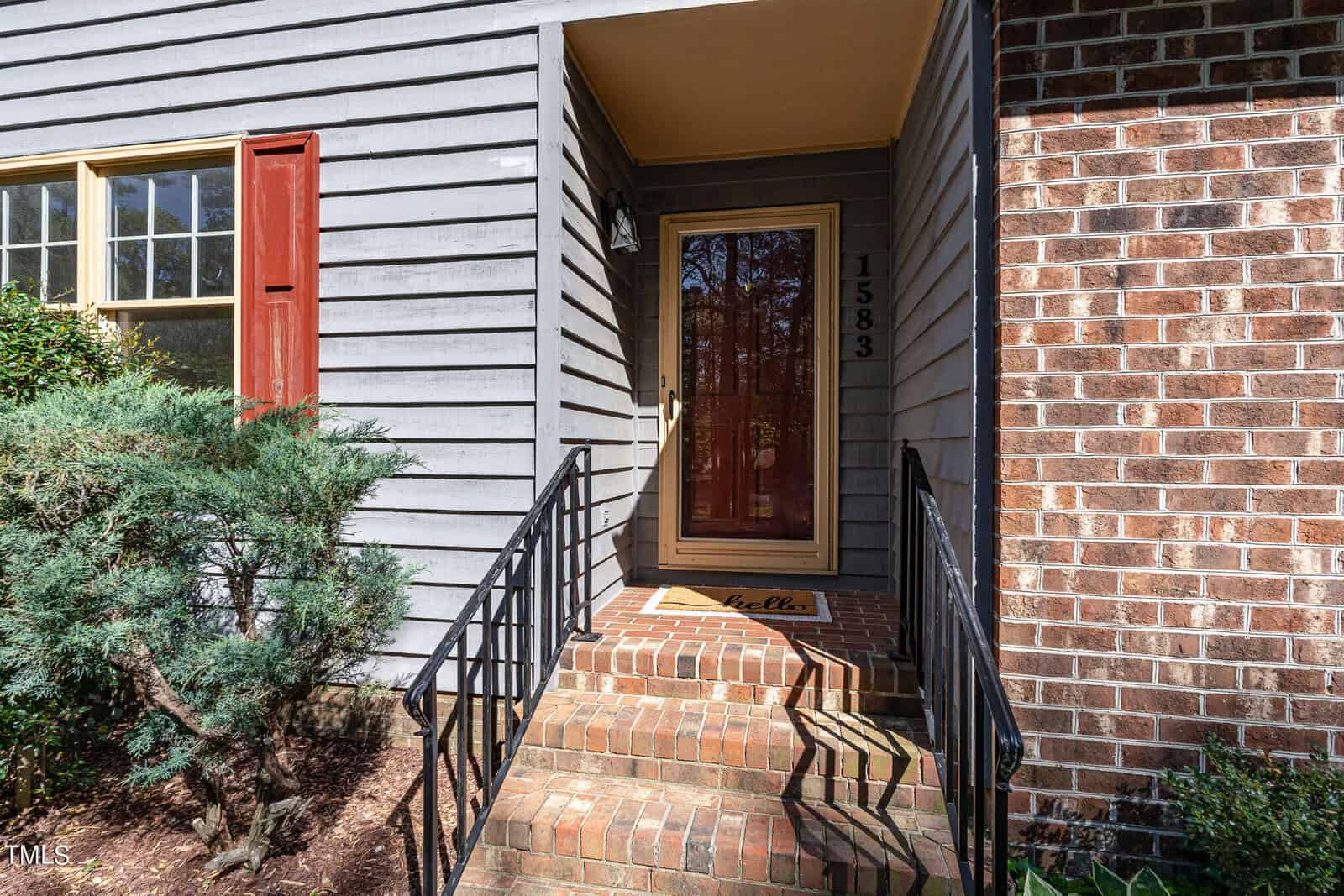 A small brick staircase with black metal railings leads to a front door with a glass center. The house has a mix of blue wooden siding and red brickwork. The house number "1583" is beside the door. A welcome mat that says "hello" is in front of the door.