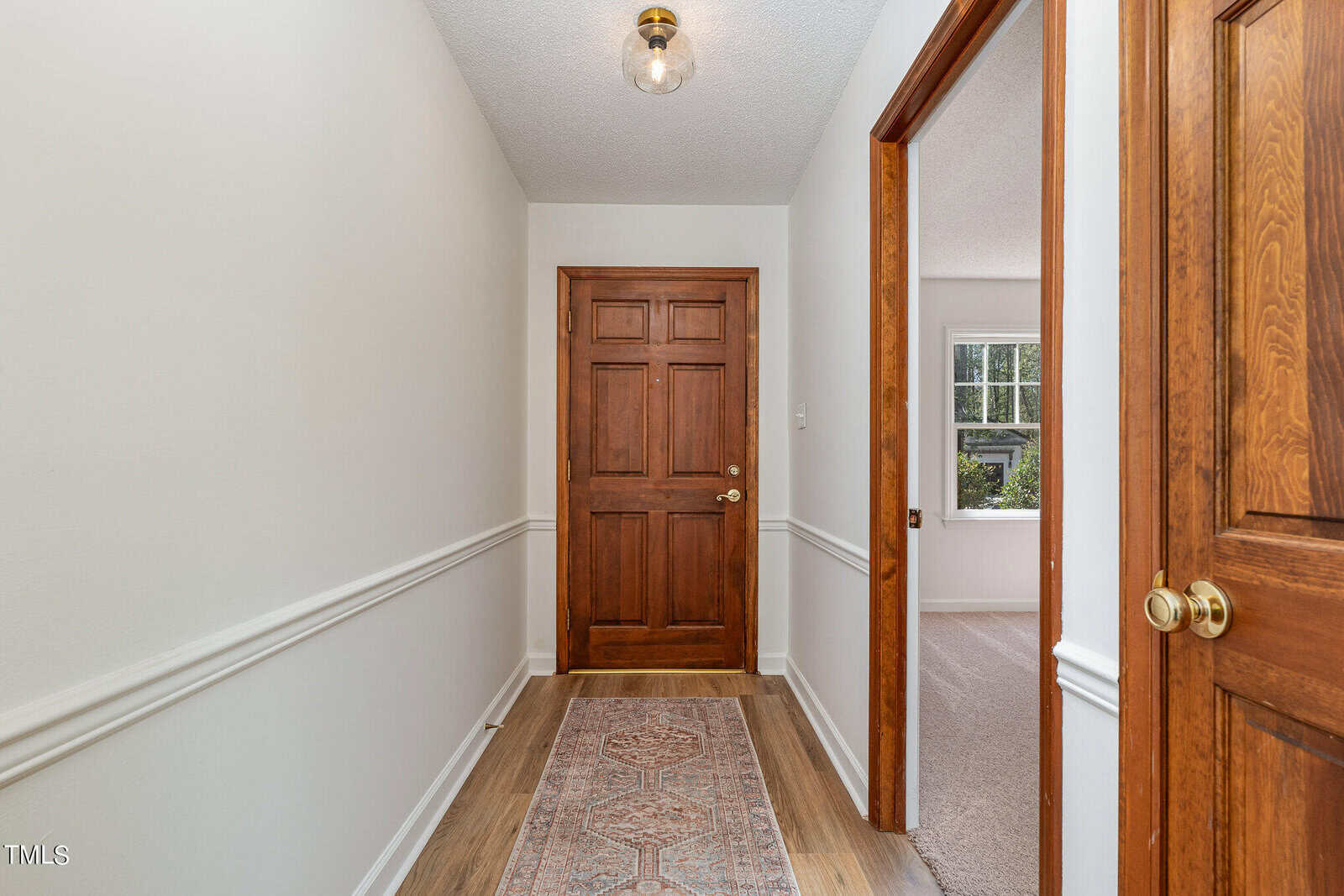 A hallway with white walls, wooden baseboards, and a light brown wood floor. A light fixture hangs from the ceiling. The hallway leads to a closed wooden door. An open door on the right reveals a room with a window. A rug runs the length of the hallway.