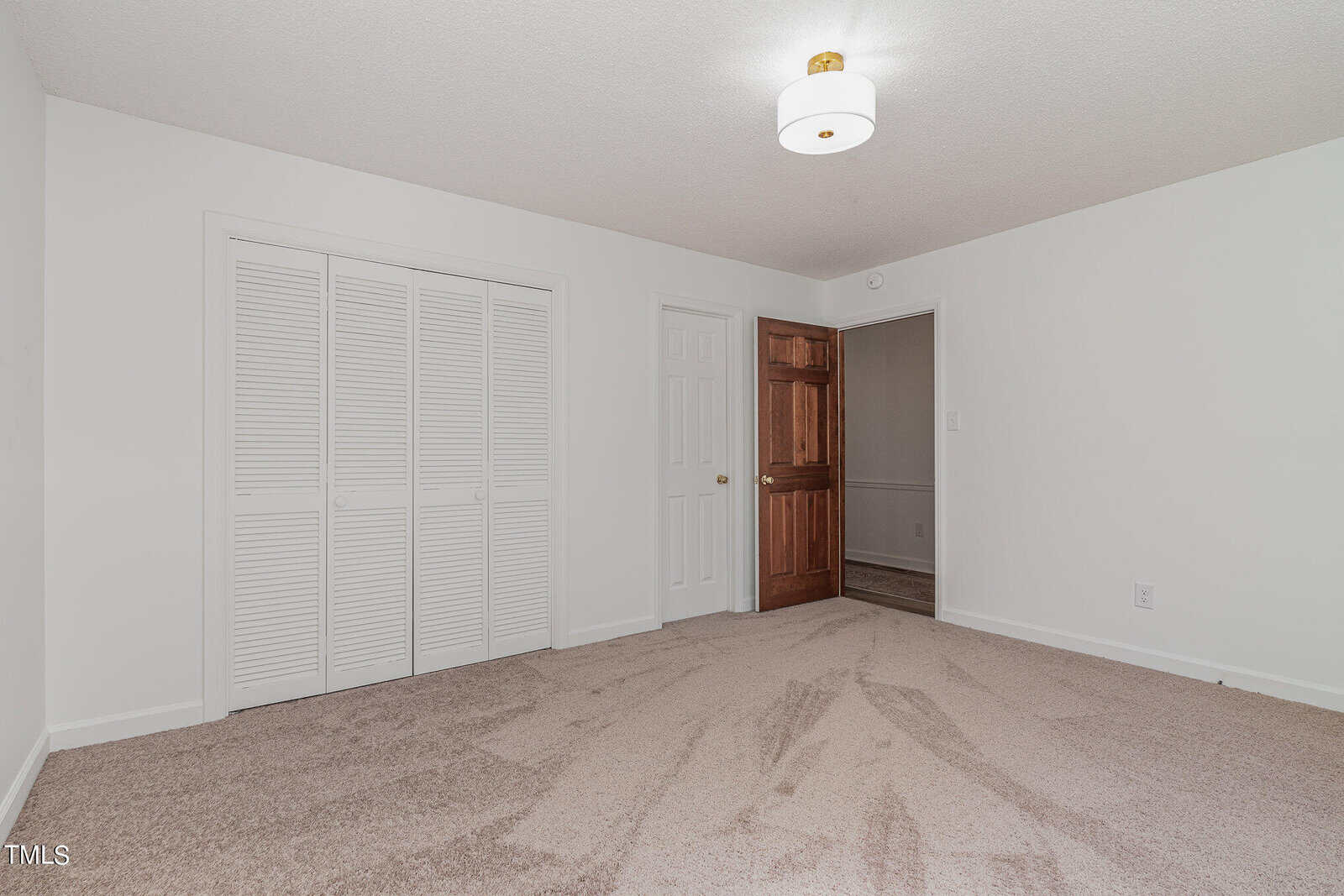 A vacant room with beige carpet, white walls, and a white ceiling. There is a closed louvered door to the left, a white door in the center, and a wooden door on the right. A modern ceiling light fixture hangs from the center of the ceiling.