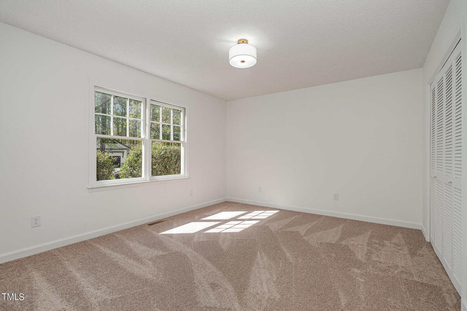 A bright and empty room with white walls and beige carpet. A circular ceiling light fixture is centered on the white ceiling. Sunlight from two windows on the left wall creates patterns on the carpet. A white door with louvered panels is on the right.