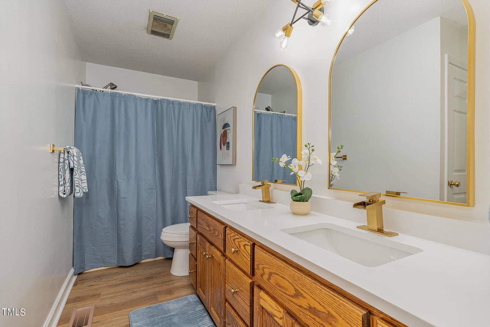 A bathroom with a double vanity featuring two white sinks and wooden cabinets. Above each sink is a large arched mirror with gold accents. The shower has a blue curtain, and there's a small painting on the wall. The floor is wooden, and the lights are on.