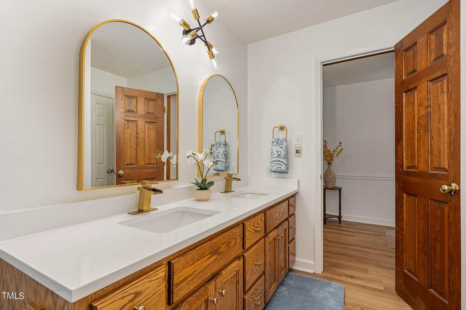 A bathroom with a double vanity featuring white countertops and wooden cabinets with gold handles. Above the sinks are two round mirrors with gold frames and wall-mounted light fixtures. A wooden door is ajar, and a small plant sits on the counter.