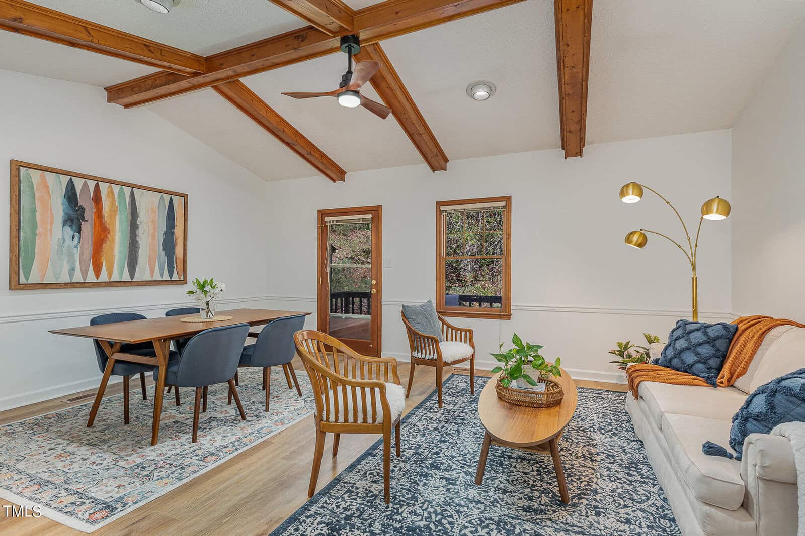A cozy living and dining area with wooden beams on the ceiling. The space features a dining table with four blue chairs, a couch, two wooden chairs, and a round coffee table. Large art pieces decorate the walls, and there are potted plants and floor lamps enhancing the ambiance.
