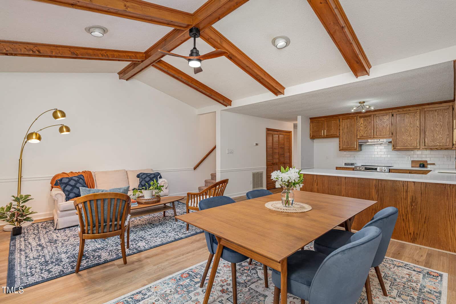 A cozy living and dining area with wooden beam ceilings. The space features a white sofa with blue cushions, wooden accent chairs, and a patterned rug. The dining area has a wooden table with blue chairs. The kitchen in the back has wooden cabinets and white countertops.