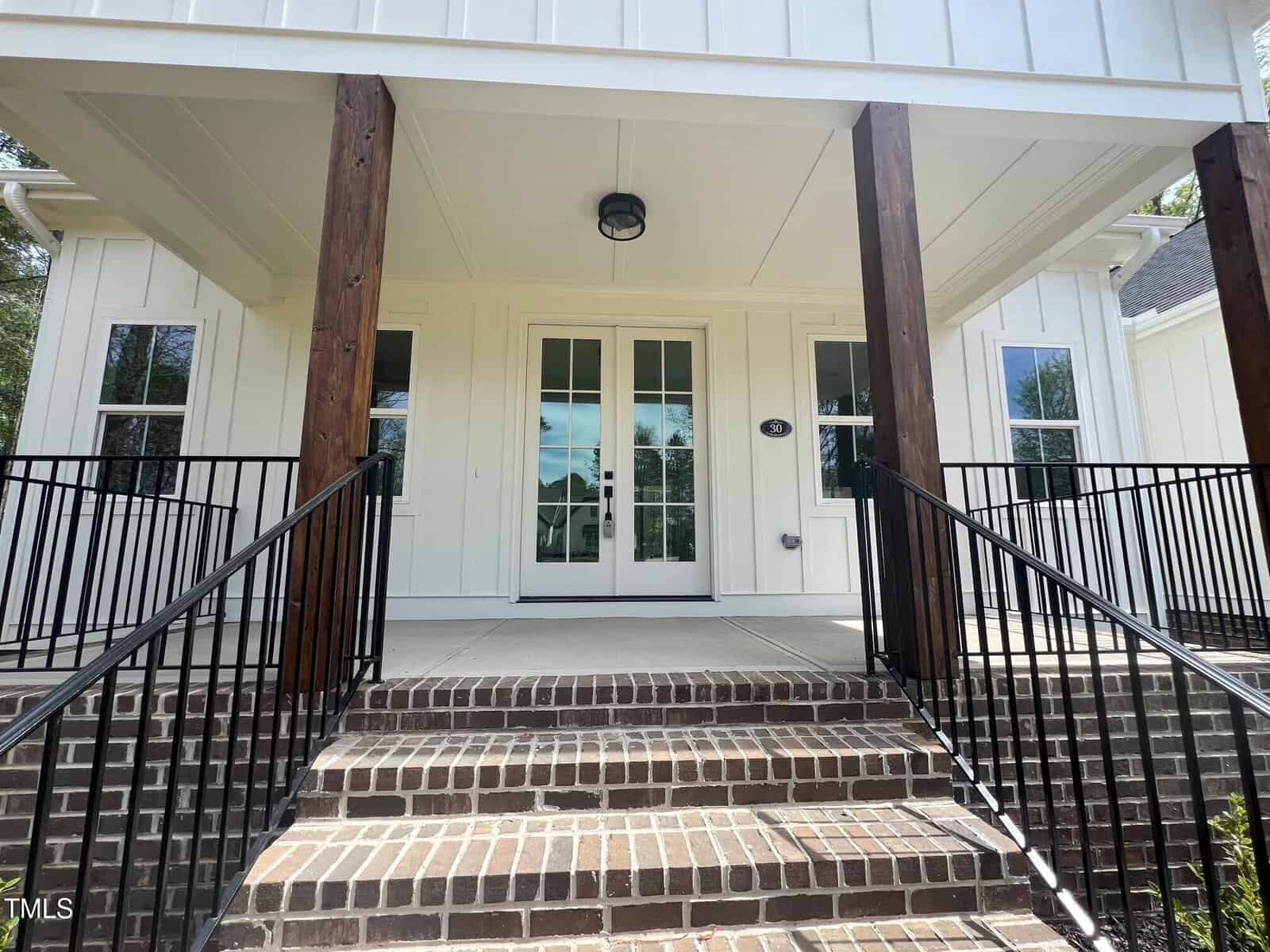 A front porch with brick steps leading to double glass doors, flanked by tall wooden columns and black metal railings. The house exterior is white with vertical paneling and windows on either side of the entrance.