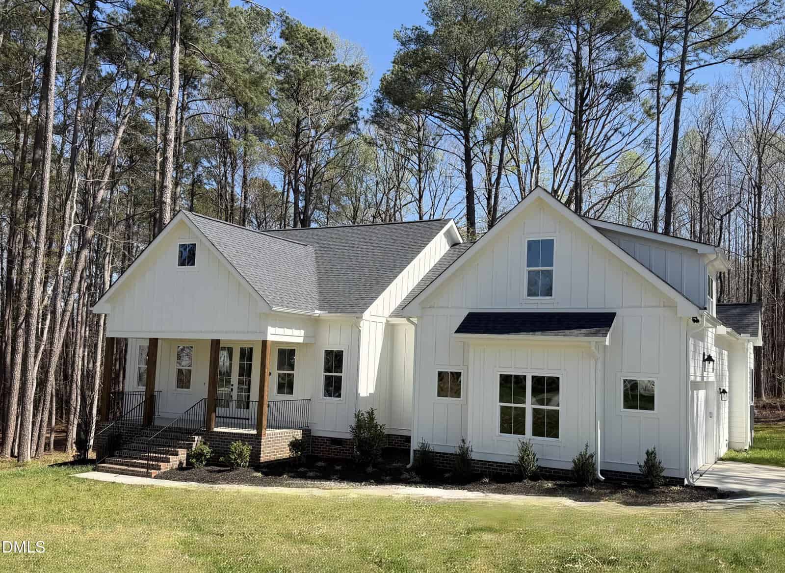A modern white house with a gray roof, front porch with steps, large windows, and black trim, surrounded by tall trees and set on a grassy lawn.