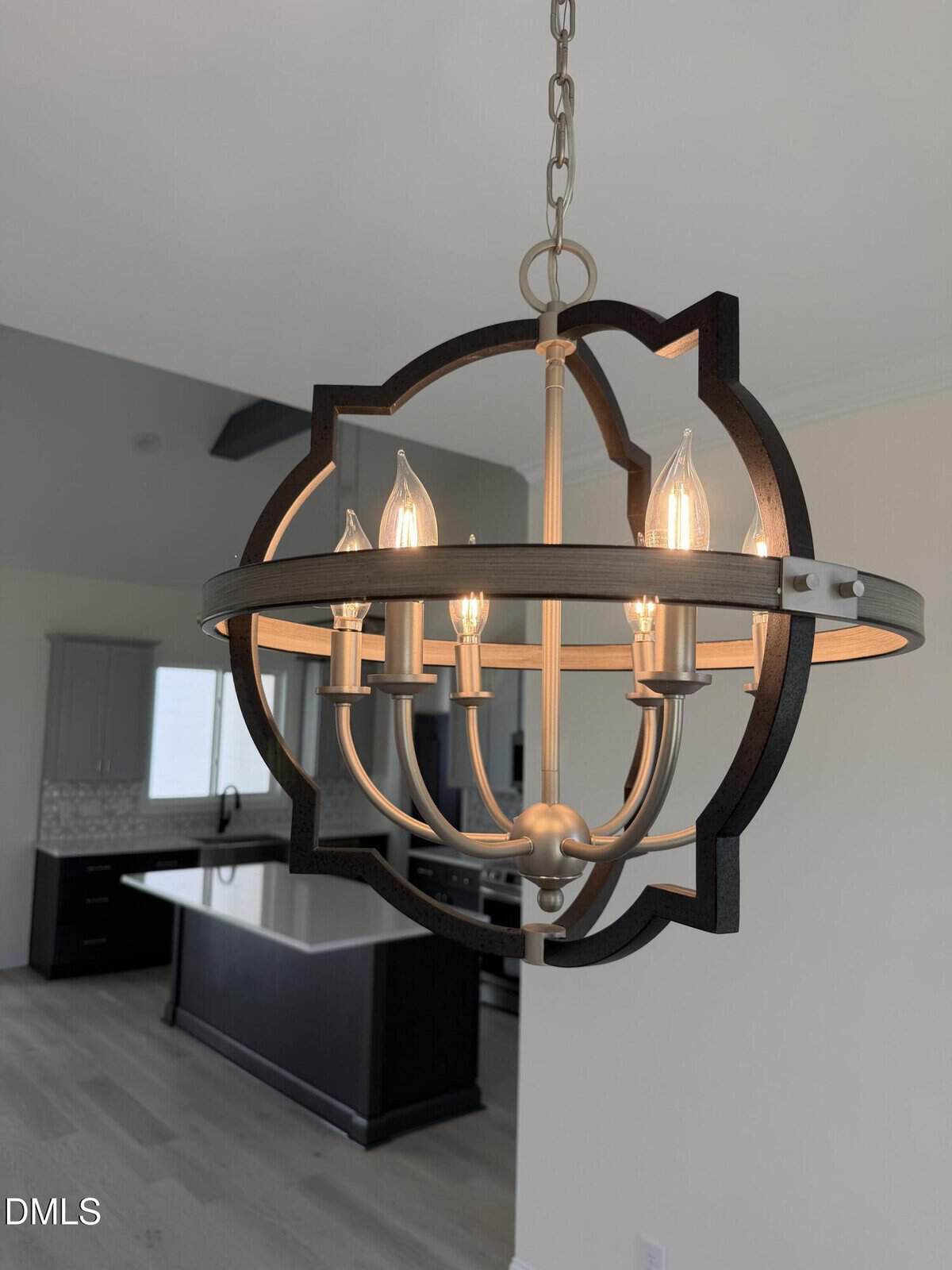 A modern chandelier with exposed bulbs hangs in a bright, open kitchen featuring light wood floors, a dark island, and white cabinetry.