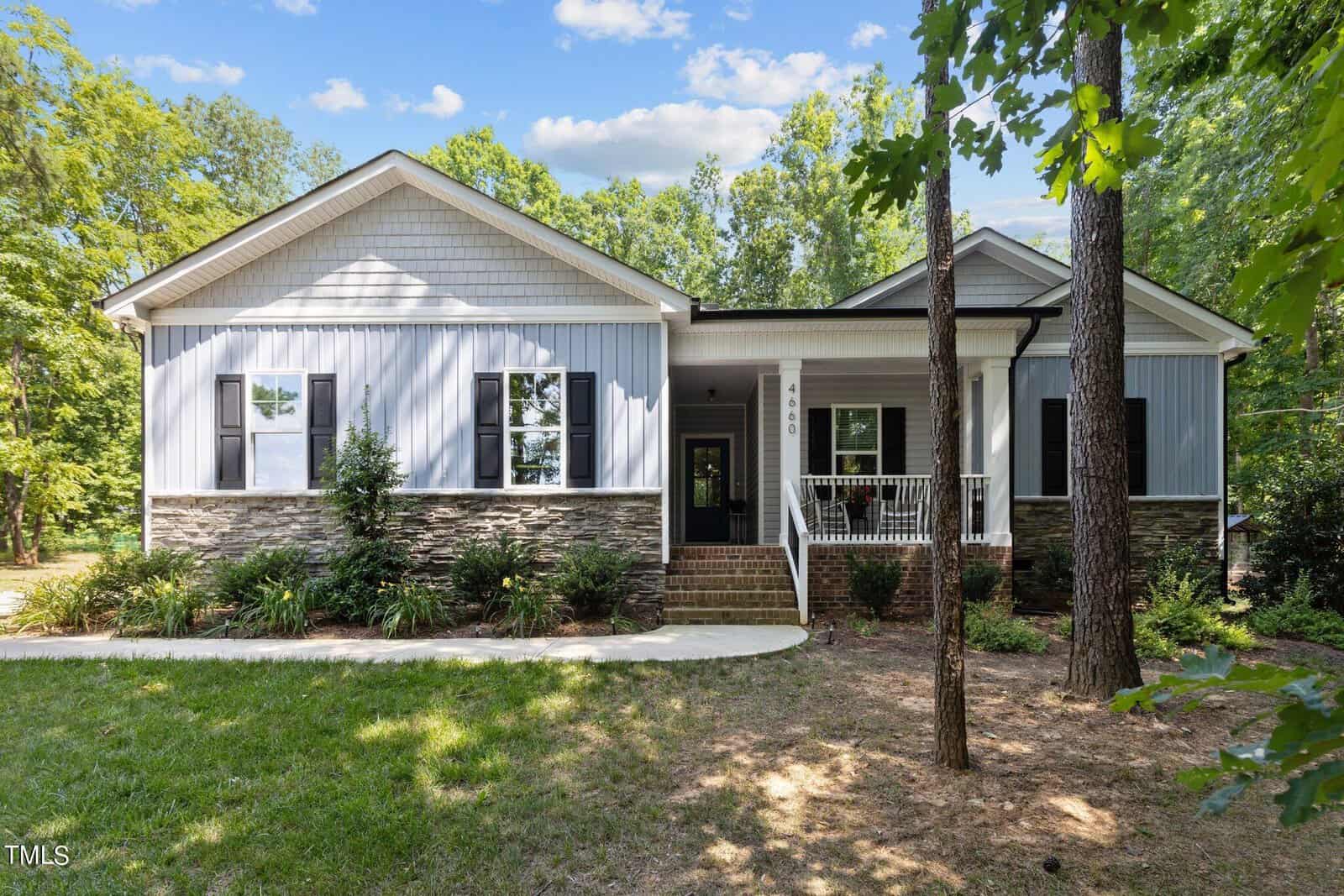 A charming single-story house on 4660 Hancock Road with a front porch, set against a backdrop of trees. The exterior features a stone and light blue siding combination, with white accents and black shutters. A well-maintained lawn and pathway lead to the entrance, echoing Oxford charm.