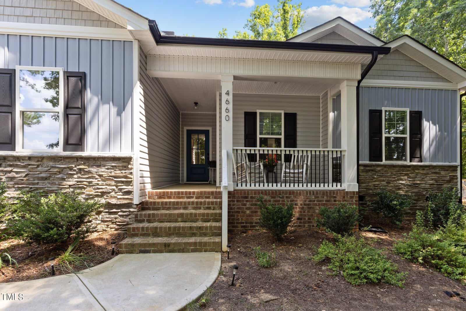 A modern house with gray siding and stone accents, featuring a small porch with a black door, white railings, and brick steps. The number 4660 is displayed on the porch column. There are small bushes in the landscaped front yard.