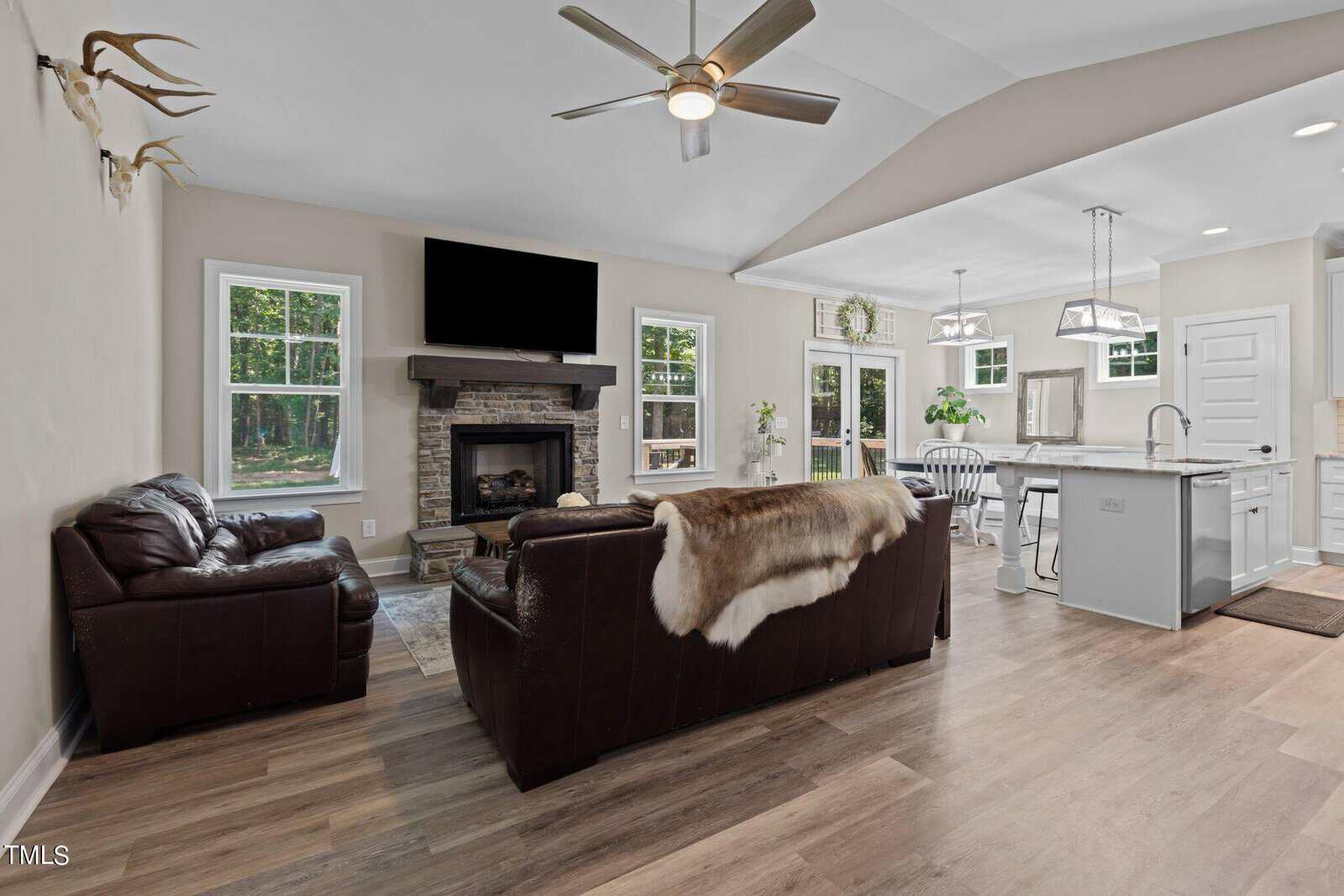 Open-concept living room and kitchen with hardwood floors. The living area features two dark leather sofas and a stone fireplace with a mounted TV. The kitchen has a large island, white cabinets, and modern appliances. A ceiling fan hangs above.