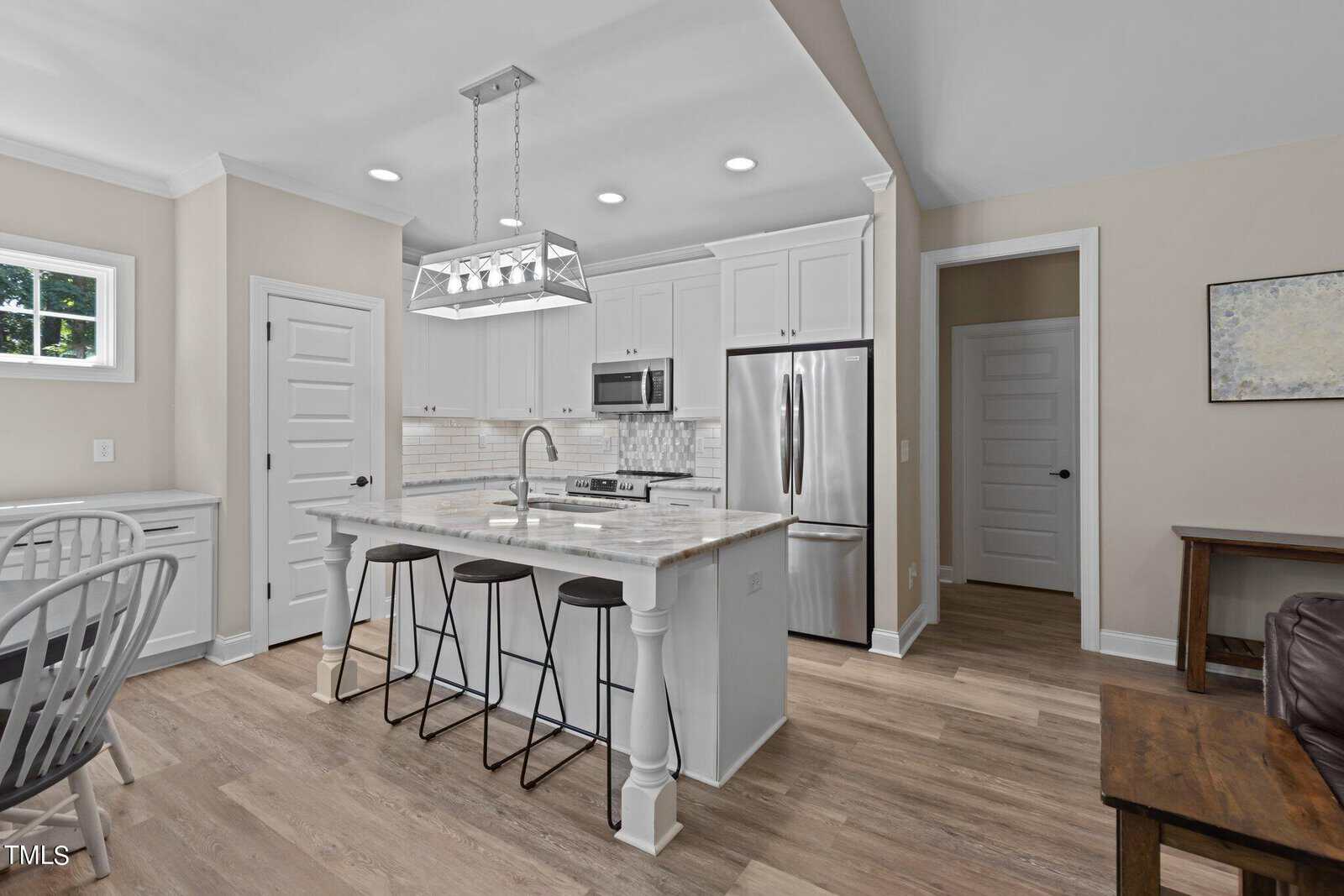 A modern kitchen with white cabinets, stainless steel appliances, and a marble-topped island. Three black stools are arranged at the island, and a geometric pendant light hangs above. Wood flooring and a soft beige wall color create a warm ambiance.