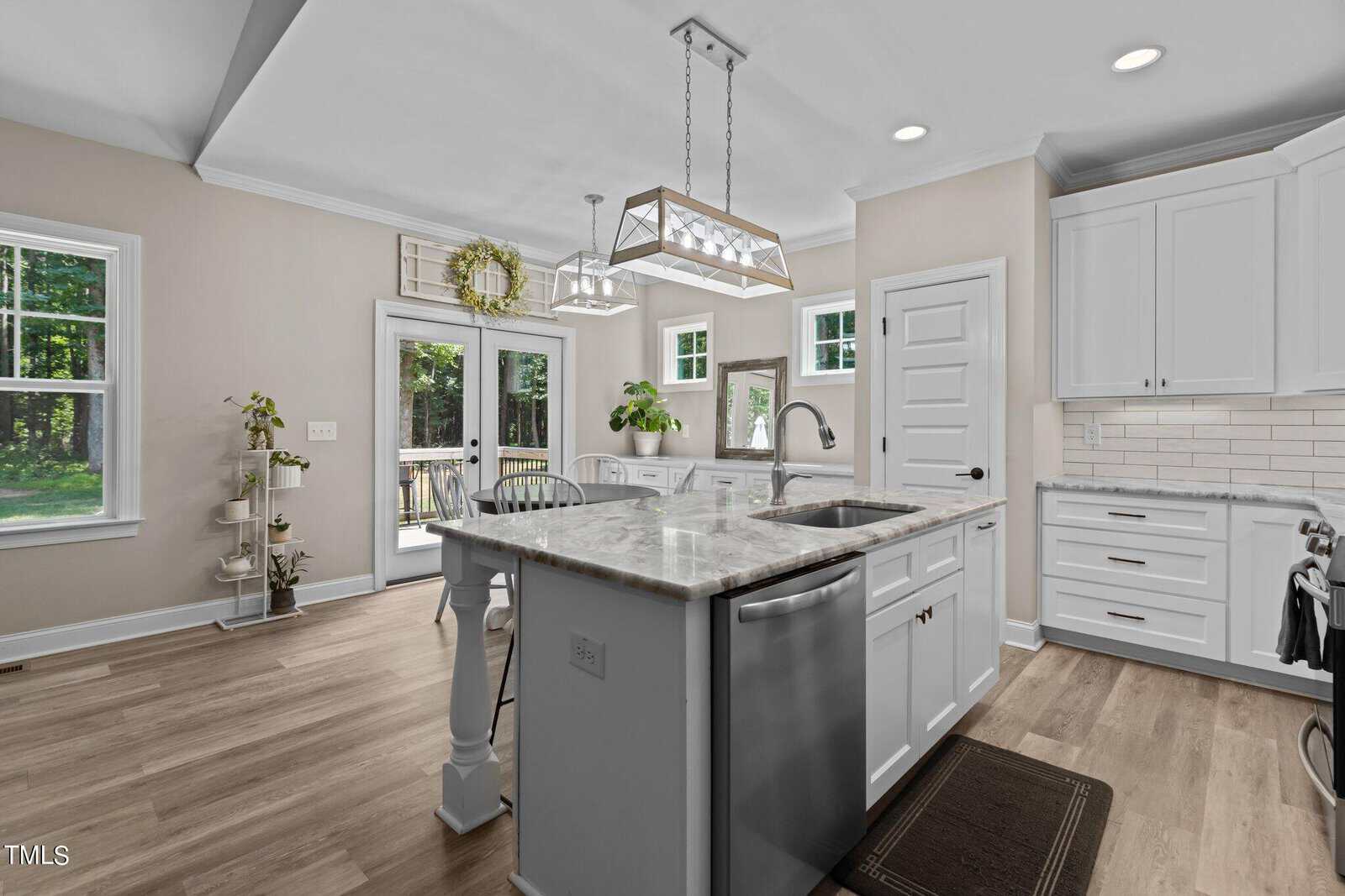 A modern kitchen featuring a central island with a sink and pendant light above. White cabinets, stainless steel dishwasher, and light wood flooring enhance the clean design. Glass doors lead to a deck with trees visible outside.