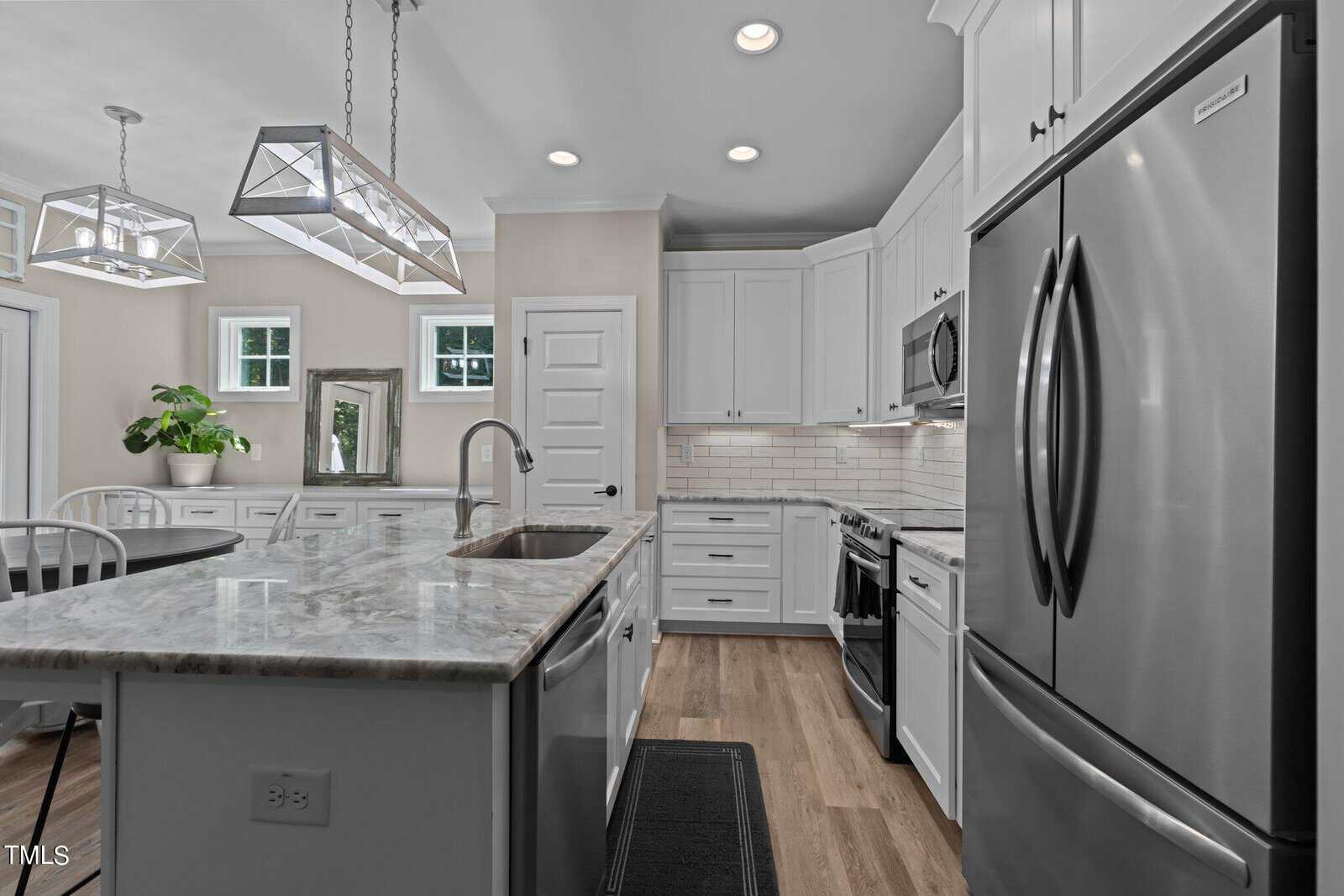 Modern kitchen with stainless steel appliances, white cabinetry, and a marble island with sink. Wooden floor, pendant lights, and a small potted plant near a window add warmth and natural light. Cozy dining area with chairs is visible in the background.