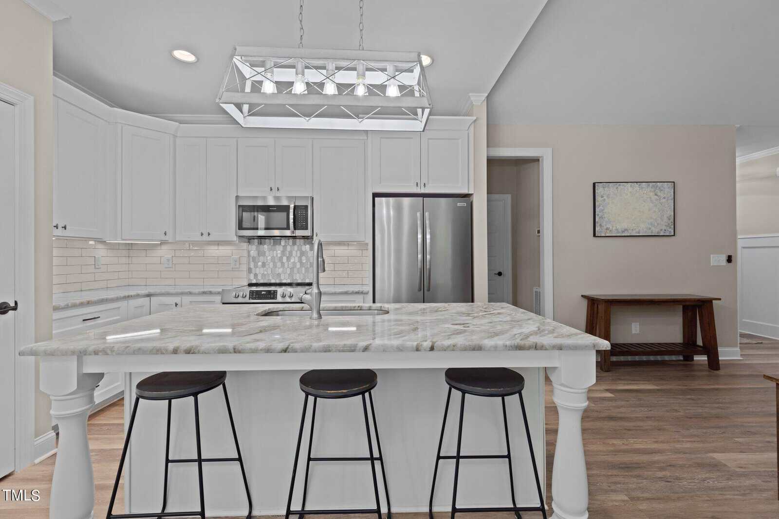 A modern kitchen featuring white cabinets, stainless steel appliances, and a marble island with three black stools. The room has wooden floors, a decorative rectangular chandelier, and a small bench along the beige wall.