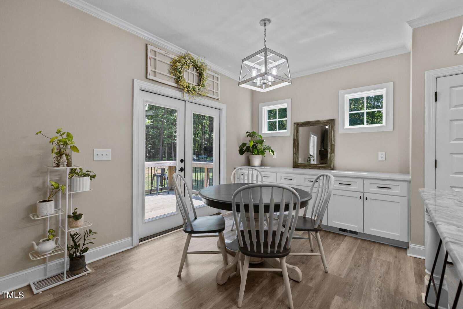 A dining room with a round black table and four gray chairs. White cabinets and a small shelf with plants are on one side. Two windows and a mirror are above the counter. Double doors lead to a deck with a view of trees. A pendant light hangs above.
