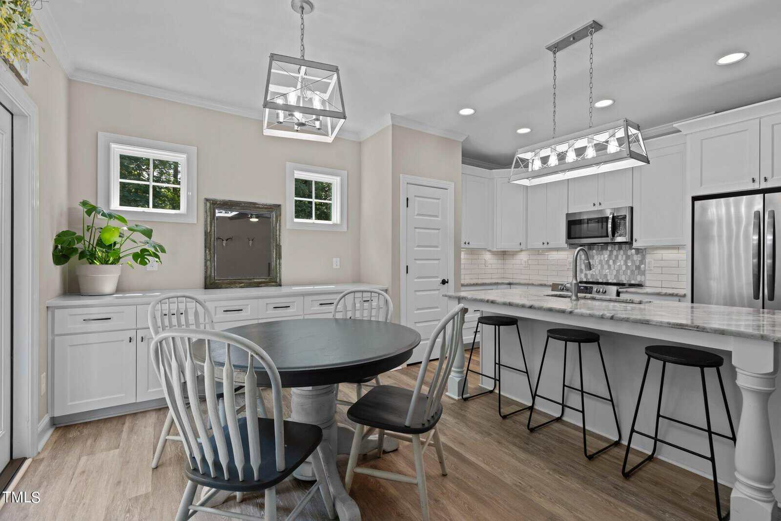 Modern kitchen with a round black dining table and white chairs. White cabinets, stainless steel appliances, and two rectangular pendant lights above a marble-topped island with black stools. A large plant is on the side counter, and light wood flooring.