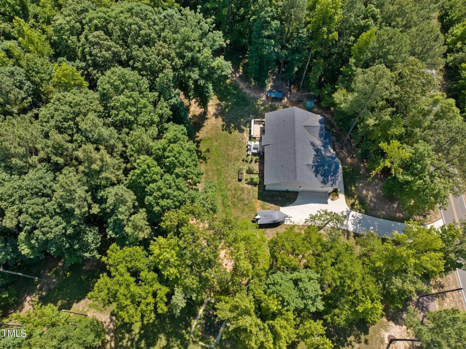 Aerial view of a house surrounded by dense greenery and trees. The light-colored house has a large roof, a driveway, and a small backyard area. Nearby, a road is partially visible through the trees.